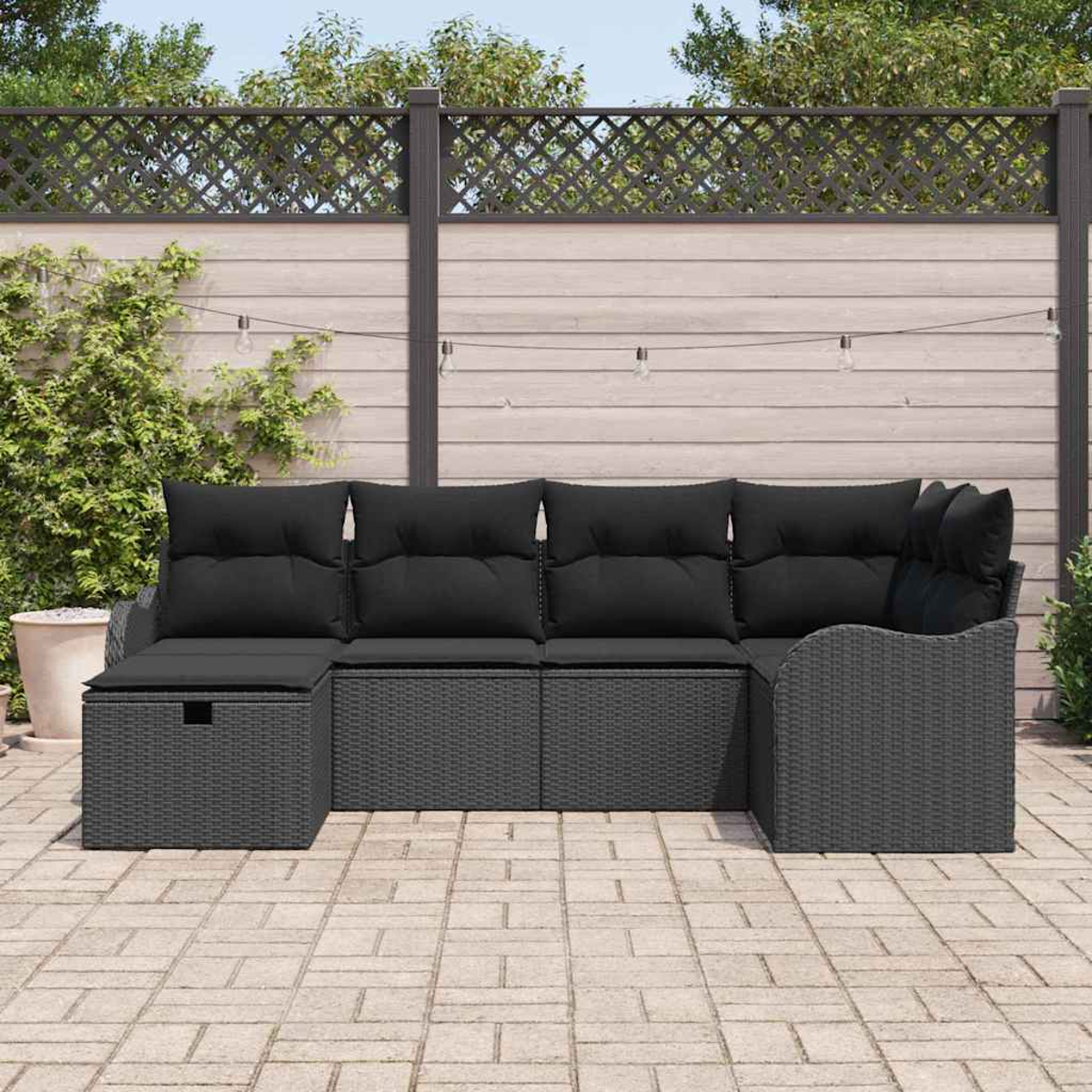 Outdoor-Ecksofa mit schwarzen Kissen. Grauer Korbrahmen. Im Hintergrund ein Holzzaun und Pflanzen.