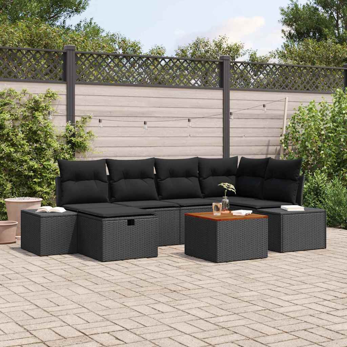 Garten-Terrassenset mit schwarzen Kissen, Holztisch und grauen Korbmöbeln. Garten mit Zaun und Pflanzen im Hintergrund.