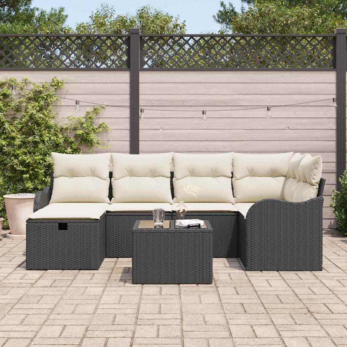 Graues Outdoor-Korbsofa mit cremefarbenen Kissen und einem kleinen Tisch. Der Hintergrund zeigt einen Holzzaun und Pflanzen.