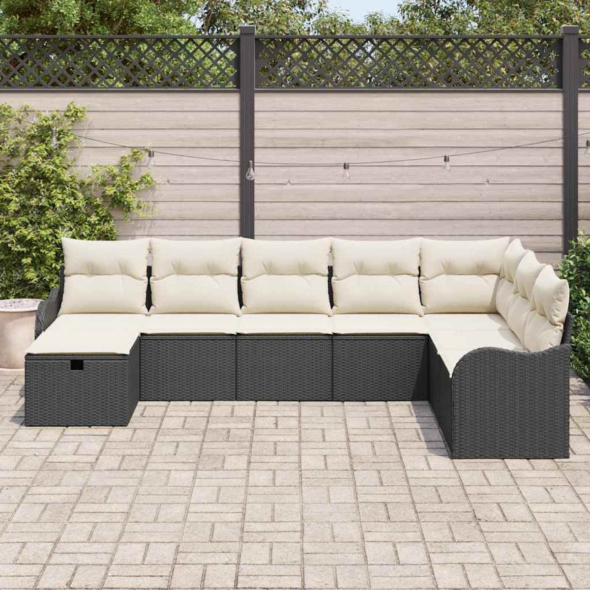 Ein Ecksofa im Freien mit cremefarbenen Kissen und dunkelgrauem Korbgeflecht. Es steht auf einer Steinterrasse mit Holzzaun im Hintergrund.