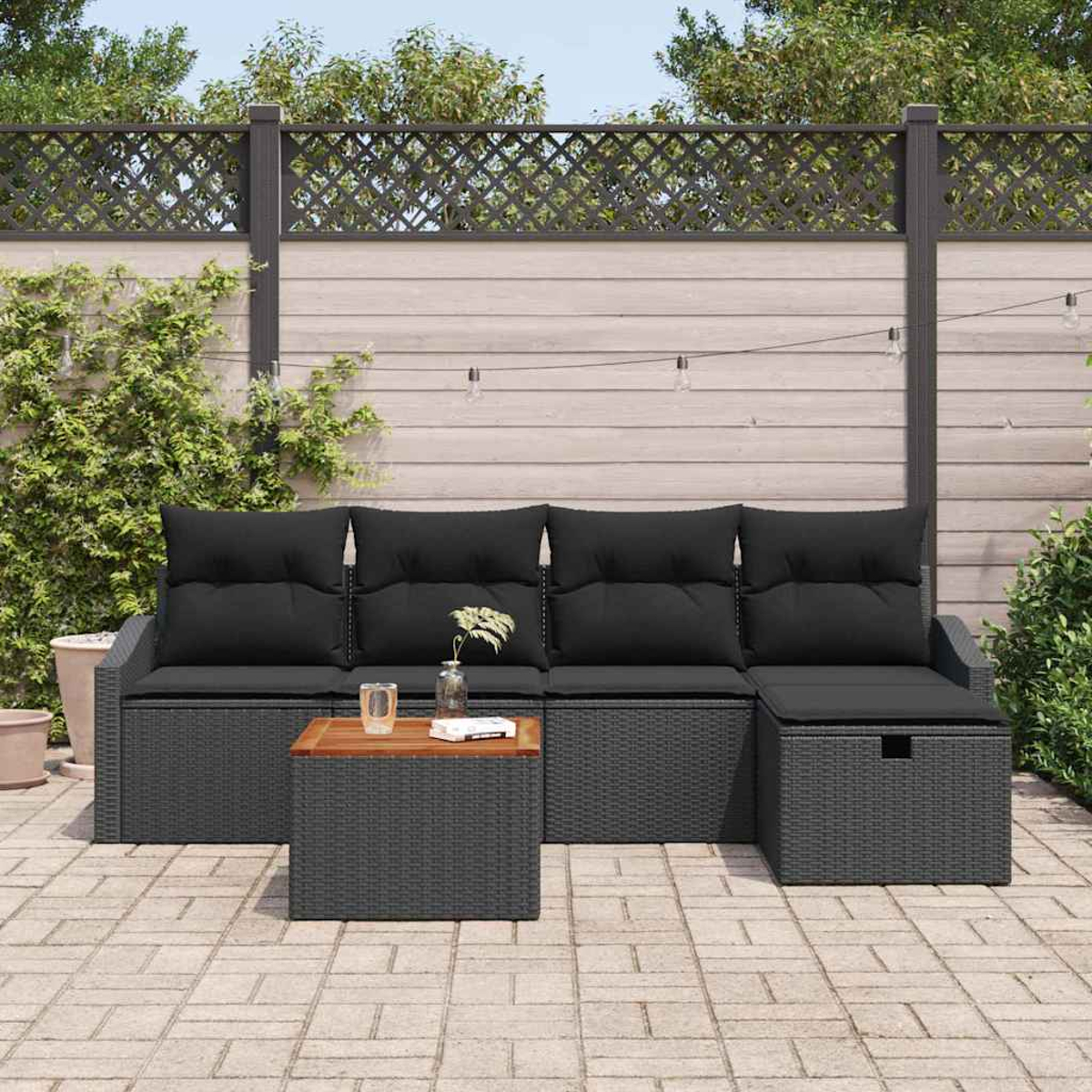 Garten-Sitzgruppe mit schwarzen Kissen. Enthält Sofa, Couchtisch und Ottomane auf einer Ziegelterrasse. Im Hintergrund ein Zaun.