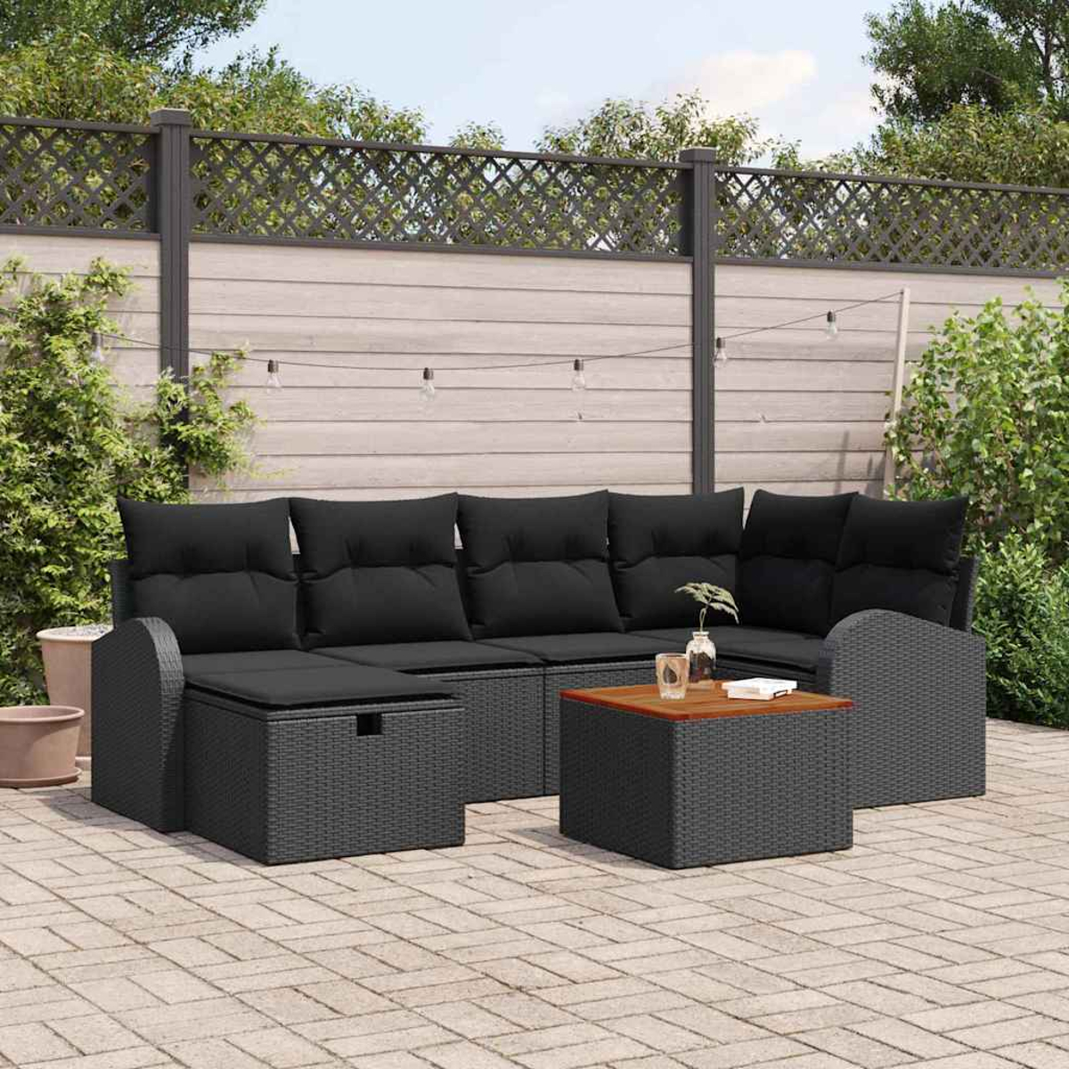 Schwarzes Outdoor-Geflechtsofa mit Kissen, kleiner Holztisch, auf einer Terrasse, mit Zaun und Pflanzen im Hintergrund.