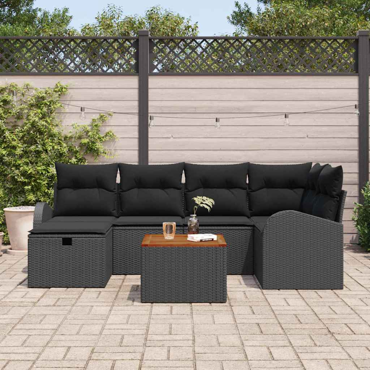 Terrassenmöbel-Set im Freien mit grauen Korbmöbeln und schwarzen Kissen. Ein Holztisch und ein dekorativer Zaun sind im Hintergrund.