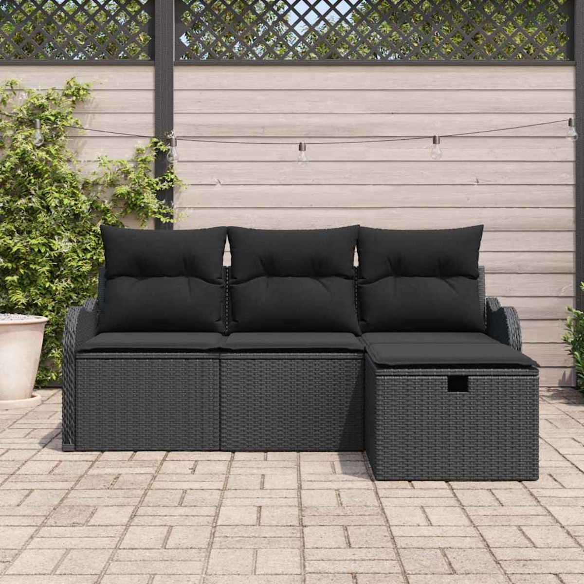 Graues Outdoor-Korbmöbel mit schwarzen Kissen. Auf einer Ziegelterrasse vor einem Holzzaun mit Kletterpflanzen. Lichterkette.