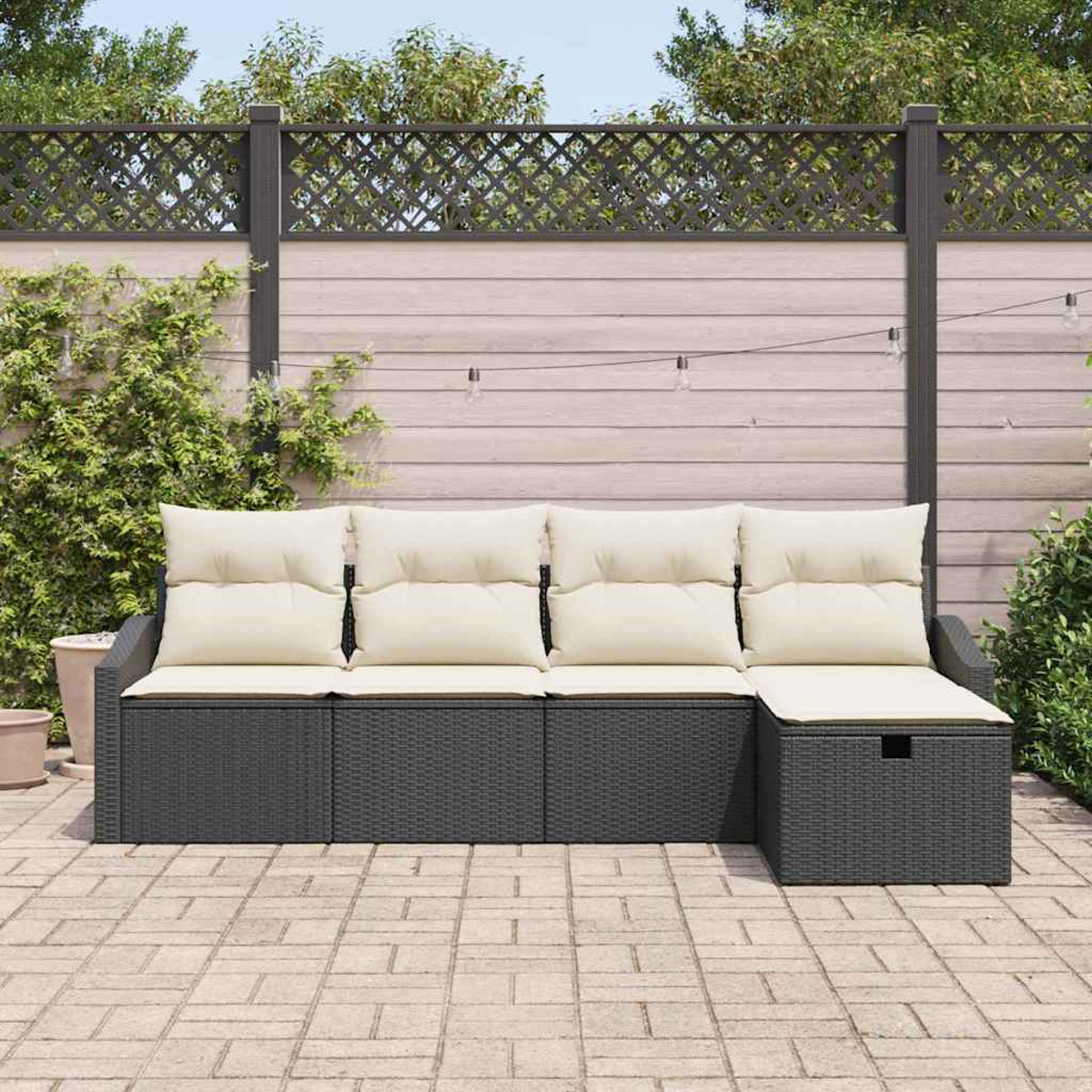 Dunkelgraues Outdoor-Sofa aus Korbgeflecht mit cremefarbenen Kissen. Es steht auf einer Ziegelterrasse. Im Hintergrund ein Holzzaun.