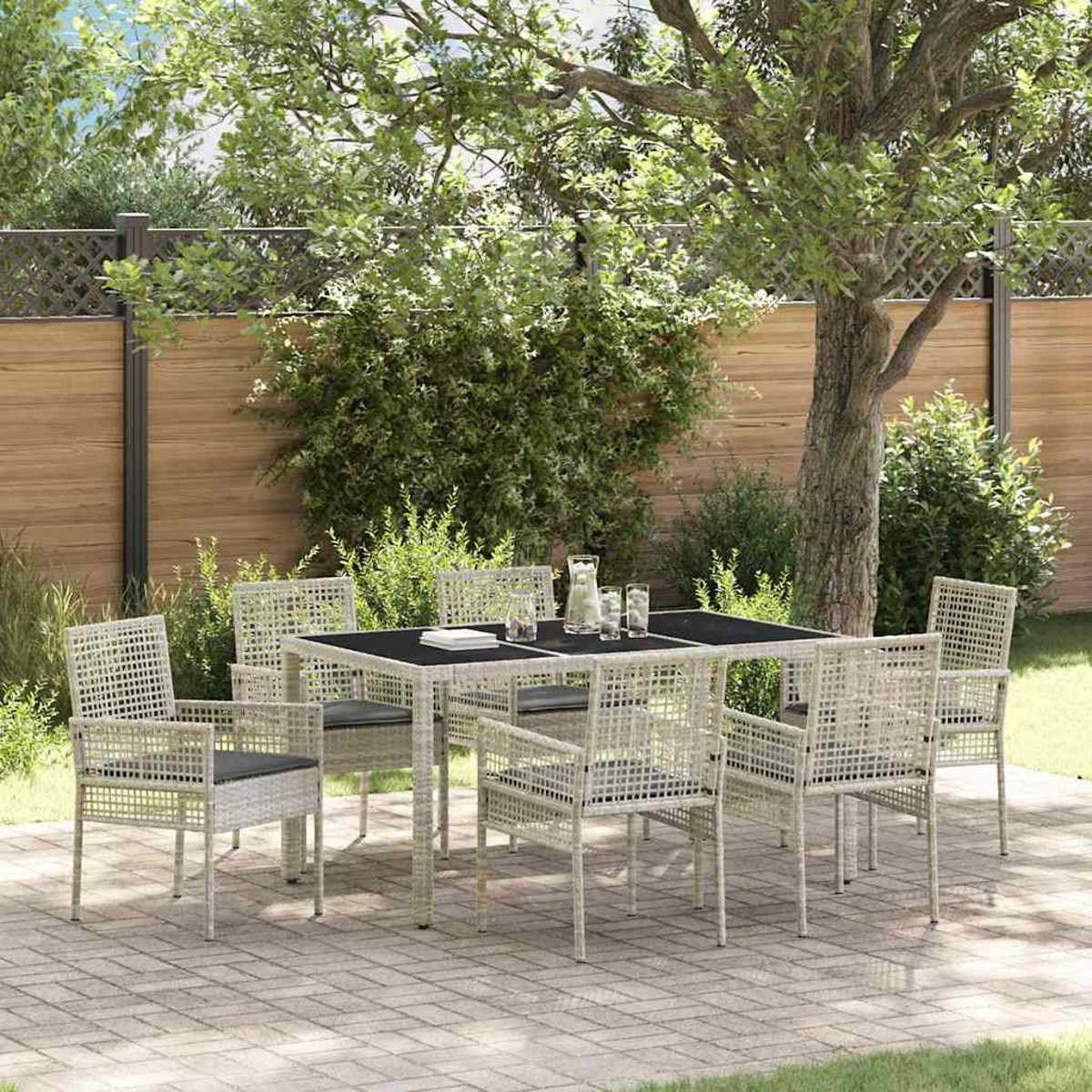 Ein weißes Terrassenset mit schwarzer Tischplatte und Kissen steht auf einem gepflasterten Bereich. Im Hintergrund sind ein Baum und ein Zaun.
