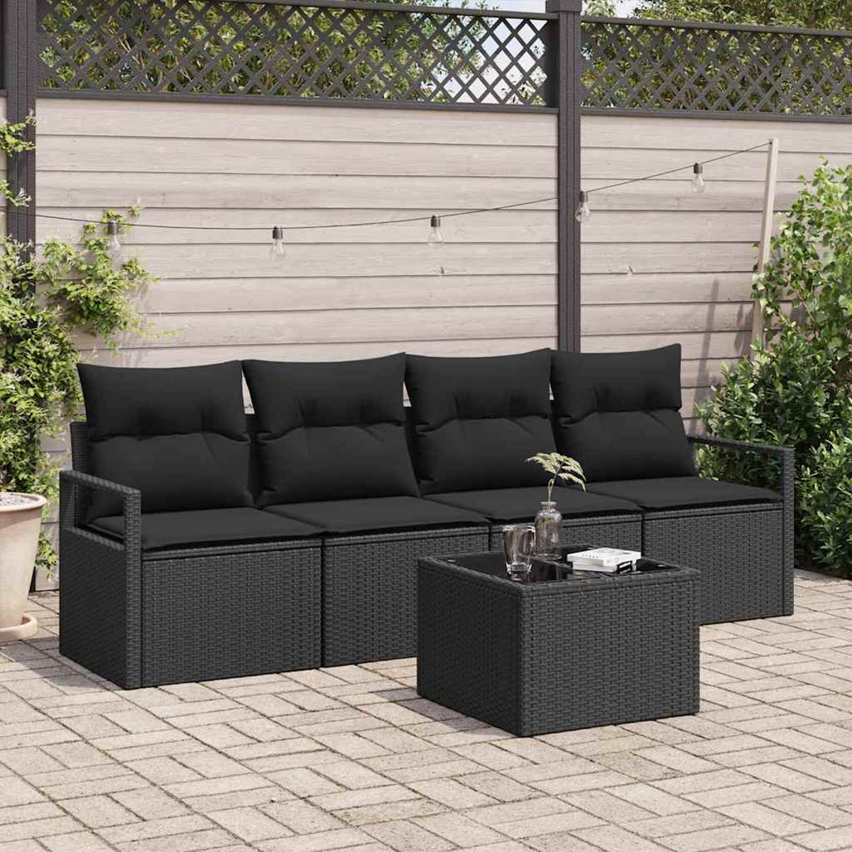 Juego de patio al aire libre. Sofá y mesa de mimbre negro con cojines negros. Iluminado por luces de cadena, contra una valla de madera y vegetación.
