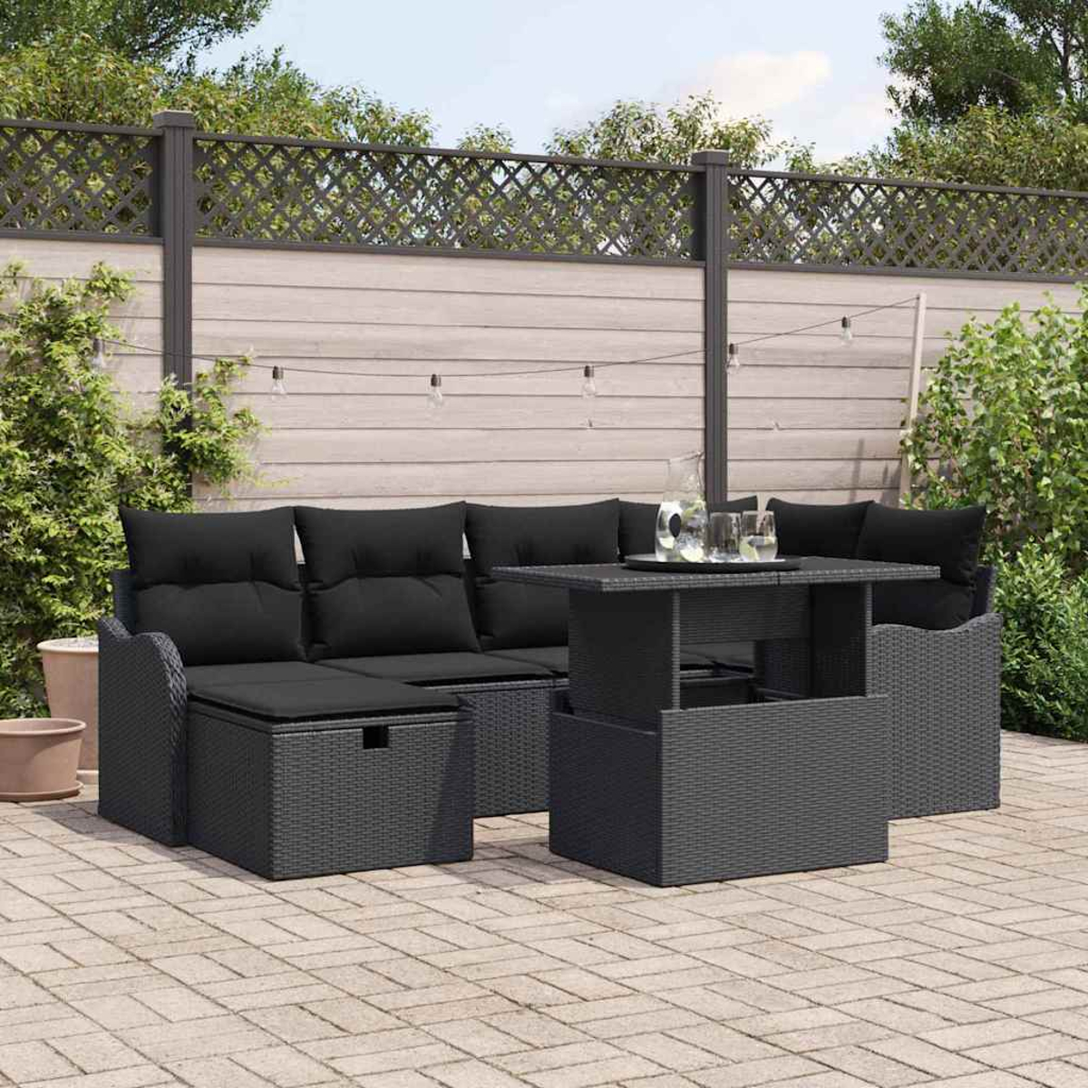 Ein schwarzes Outdoor-Wicker-Möbelset mit Kissen, Tisch und Ottomane auf einer Backstein-Terrasse. Im Hintergrund ein Zaun und Grün.