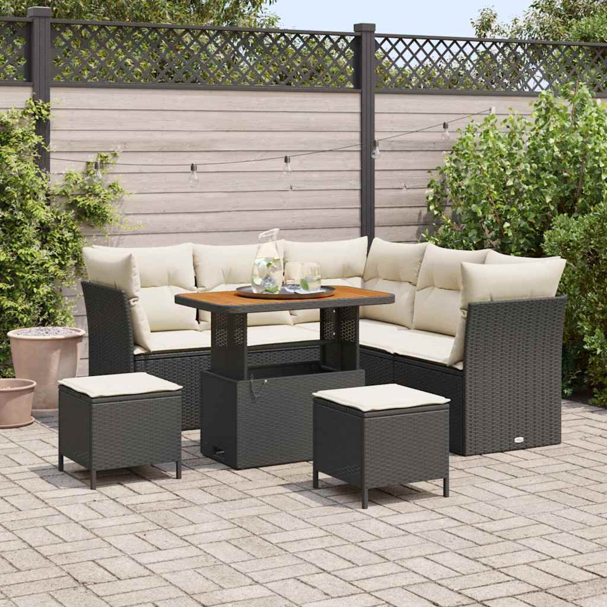 Terrassenmöbel-Set mit grauen Korbmöbeln, beigen Kissen und einem Holztisch. Zwei Hocker und ein Ecksofa sind sichtbar. Hintergrund: Zaun und Pflanzen.