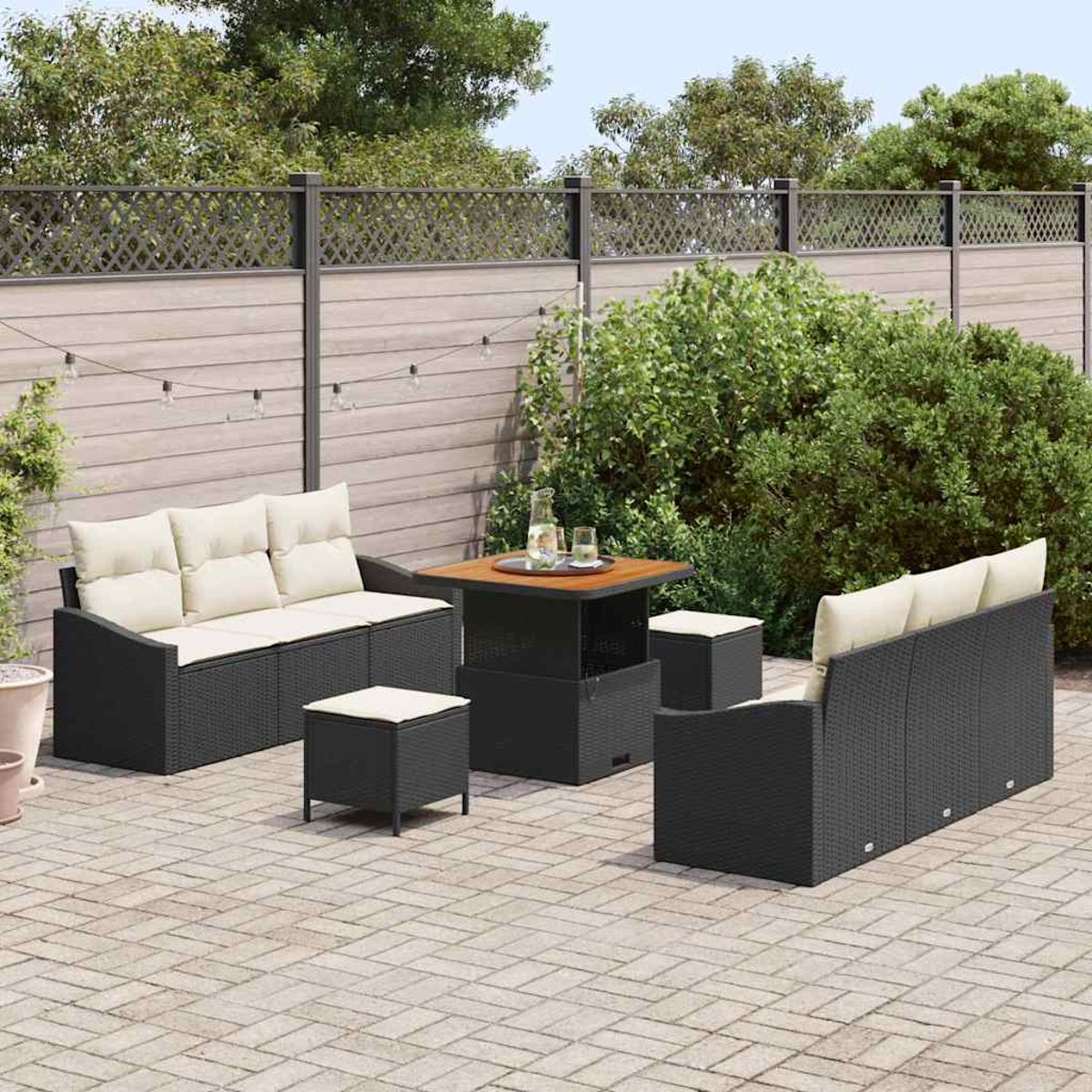 Set patio esterno: divani in vimini neri, pouf e un tavolo con cuscini beige, disposti su un patio in mattoni, con verde sullo sfondo.
