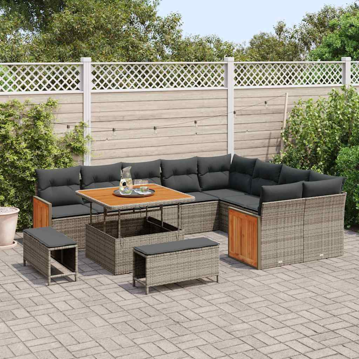 Gartenmöbel-Set. Ecksofa aus grauem Korbgeflecht mit schwarzen Kissen, Holztisch, Bänken und weißem Zaun im Hintergrund.