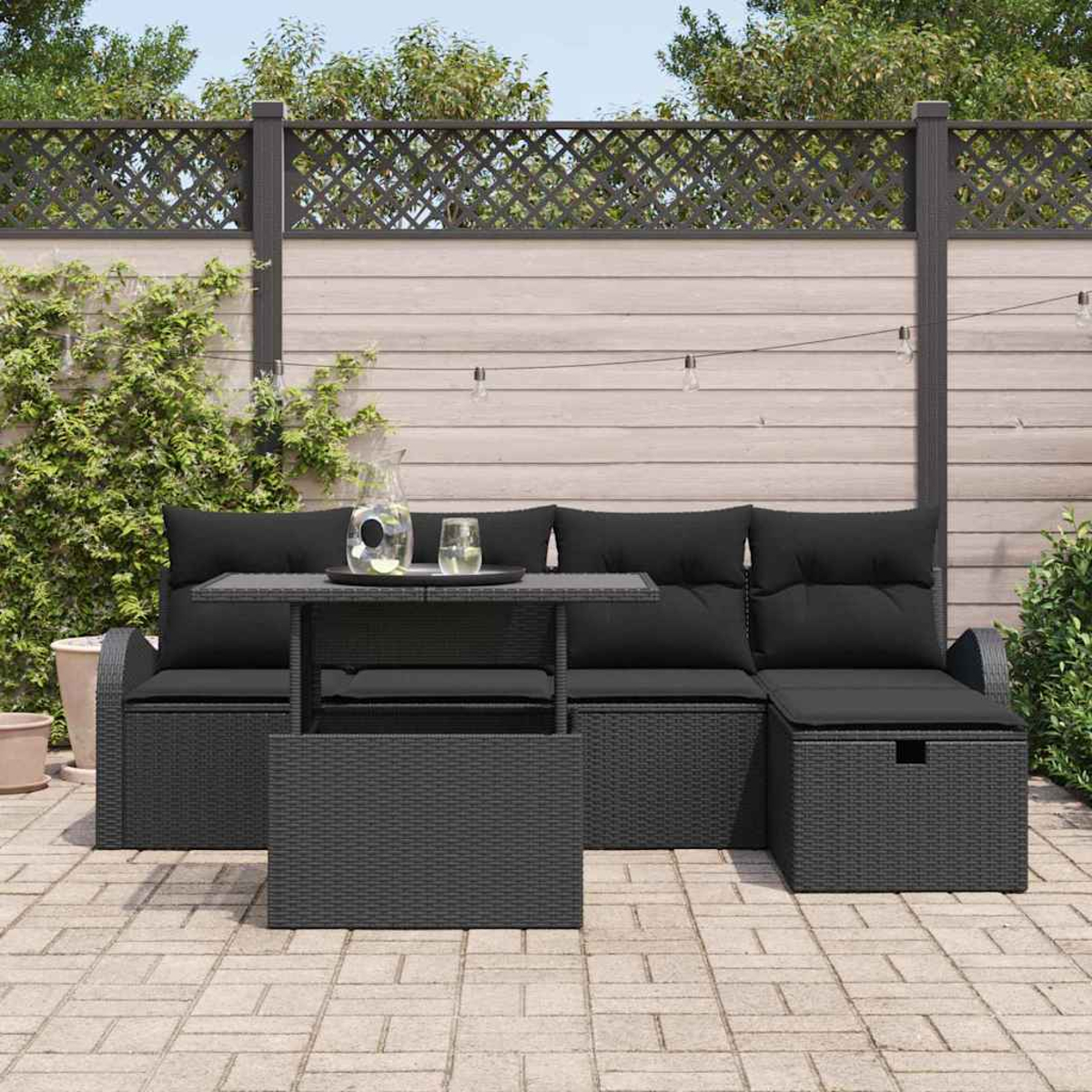 Juego de sofá y mesa de mimbre negro para exteriores en un patio. Fondo: valla de madera, vegetación y luces de cadena.