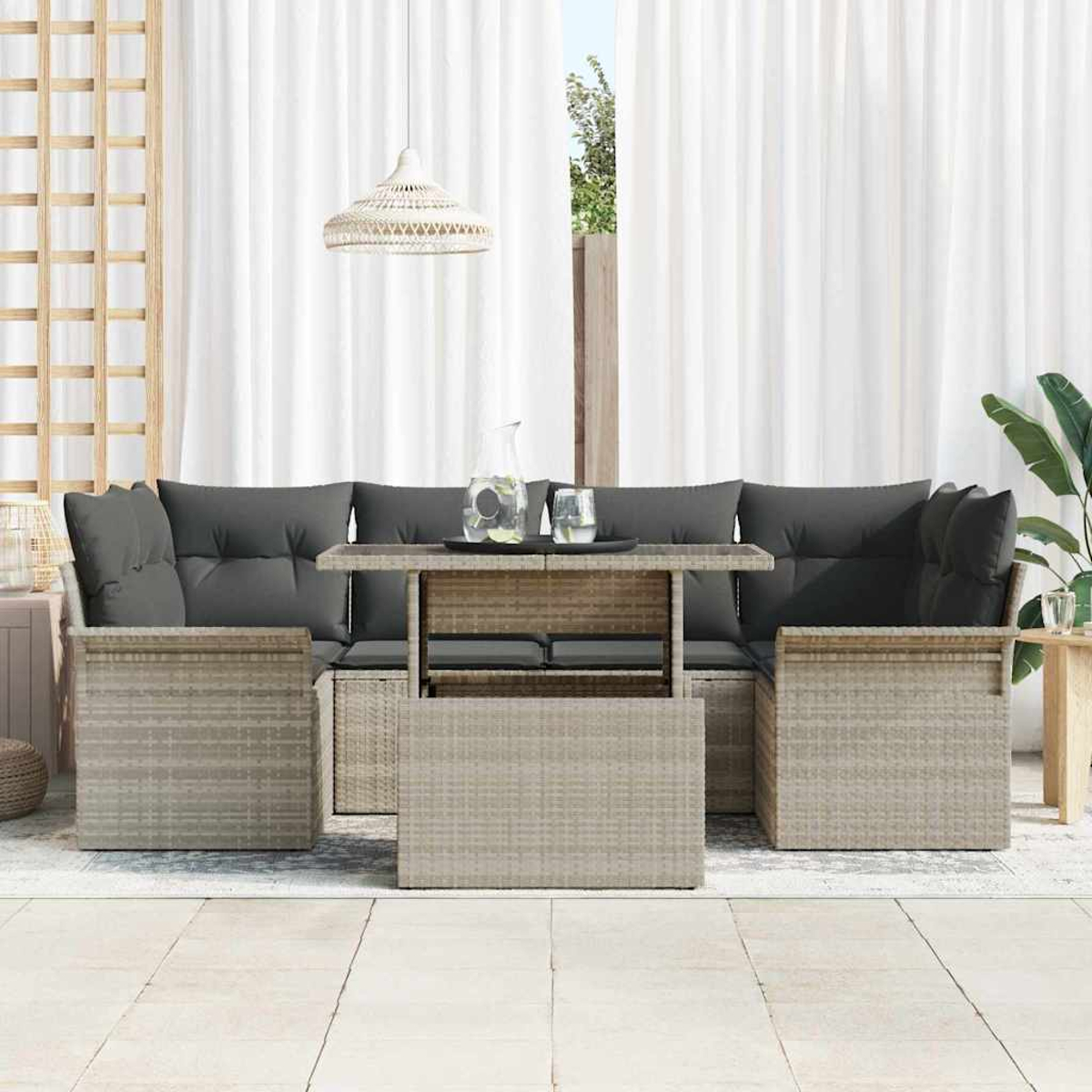 Juego de muebles de ratán para exteriores con cojines grises, mesa elevable y cortinas blancas al fondo.