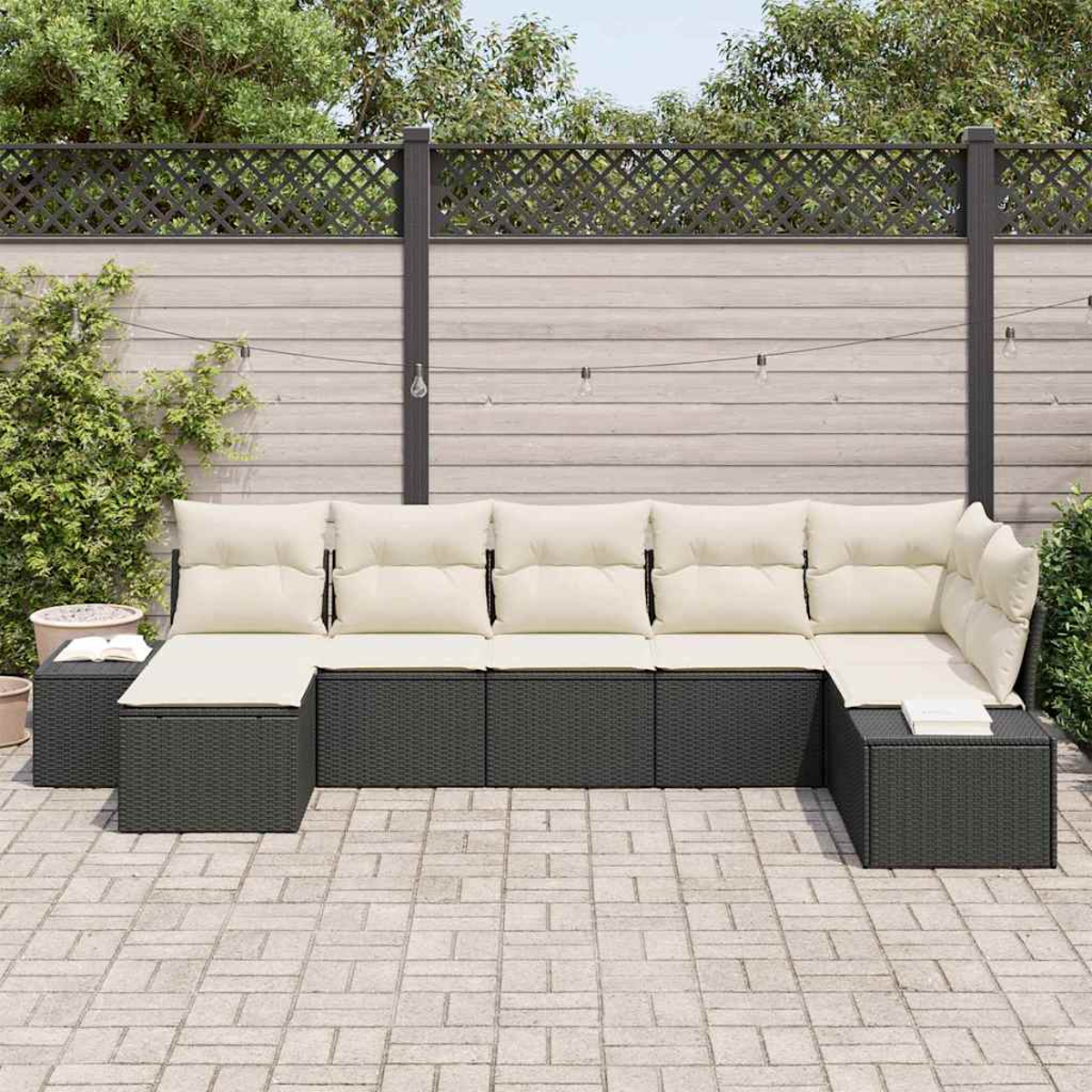 Ein Ecksofa im Freien mit cremefarbenen Kissen steht auf einer Terrasse. Ein Holzzaun, Pflanzen und Lichterketten sind im Hintergrund.