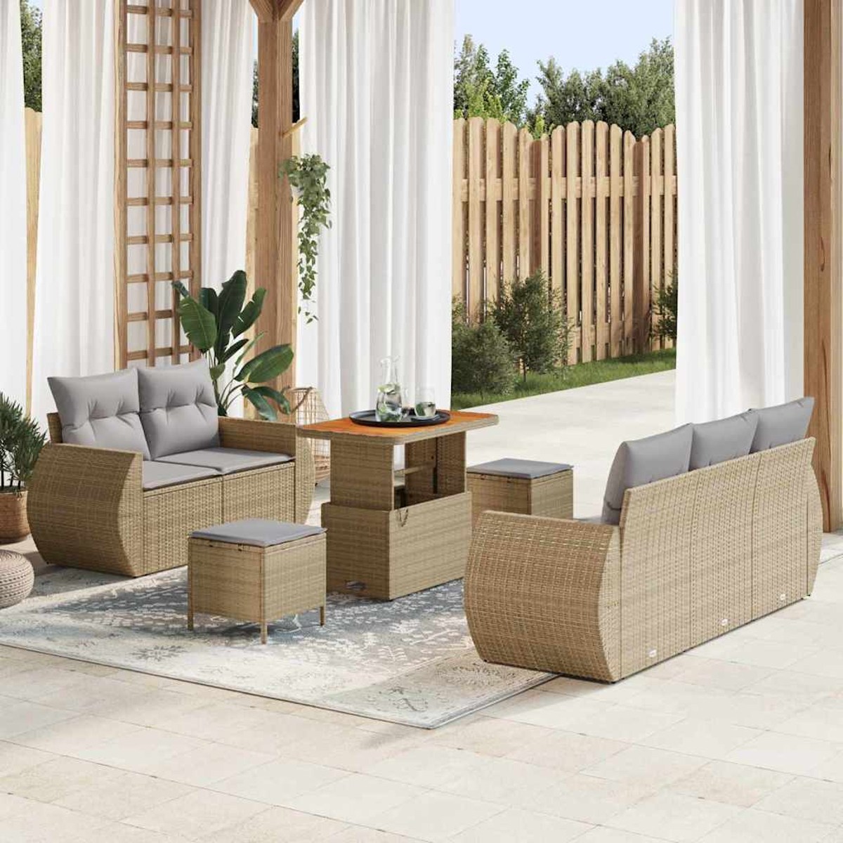 Conjunto de patio exterior con muebles tejidos, cojines grises, una mesa y taburetes. Cortinas y una valla de madera al fondo.