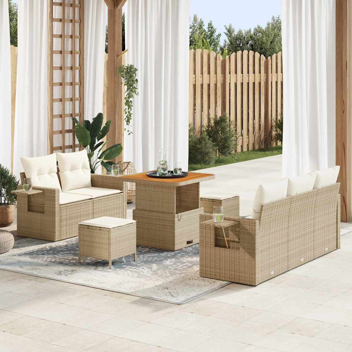 Terrassengarnitur mit beigen Korbmöbeln, weißen Kissen, Couchtisch, Teppich und weißen Vorhängen. Holzzaun im Hintergrund.