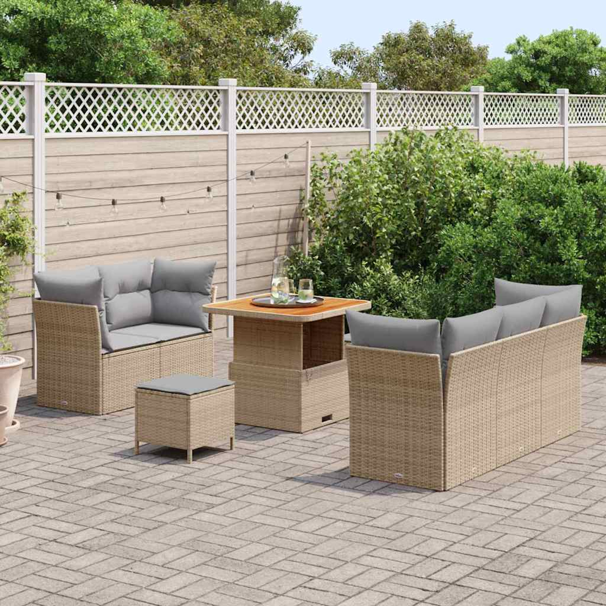 Szene im Freien mit Korbmöbeln. Auf einer gepflasterten Terrasse stehen ein Sofa, zwei Sessel, ein Tisch und ein Hocker. Im Hintergrund ein Zaun und Grün.