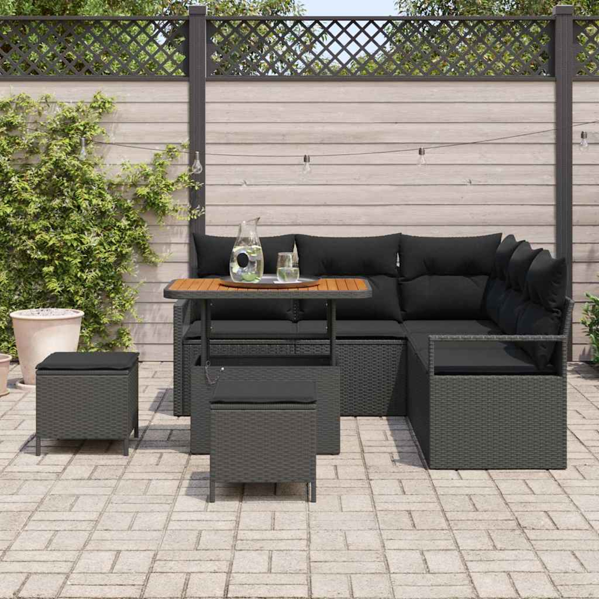 Outdoor-Ecksofa mit schwarzen Kissen, einem kleinen Tisch mit einem Krug und zwei Hockern auf einer Ziegelterrasse. Holzzaun im Hintergrund.