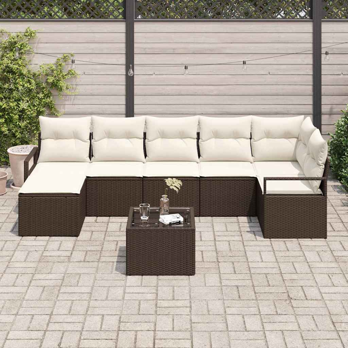 Braunes Outdoor-Korbssofa mit cremefarbenen Kissen, kleiner Tisch mit Glas und Pflanzen. Auf Steinterrasse mit Holzzaun.