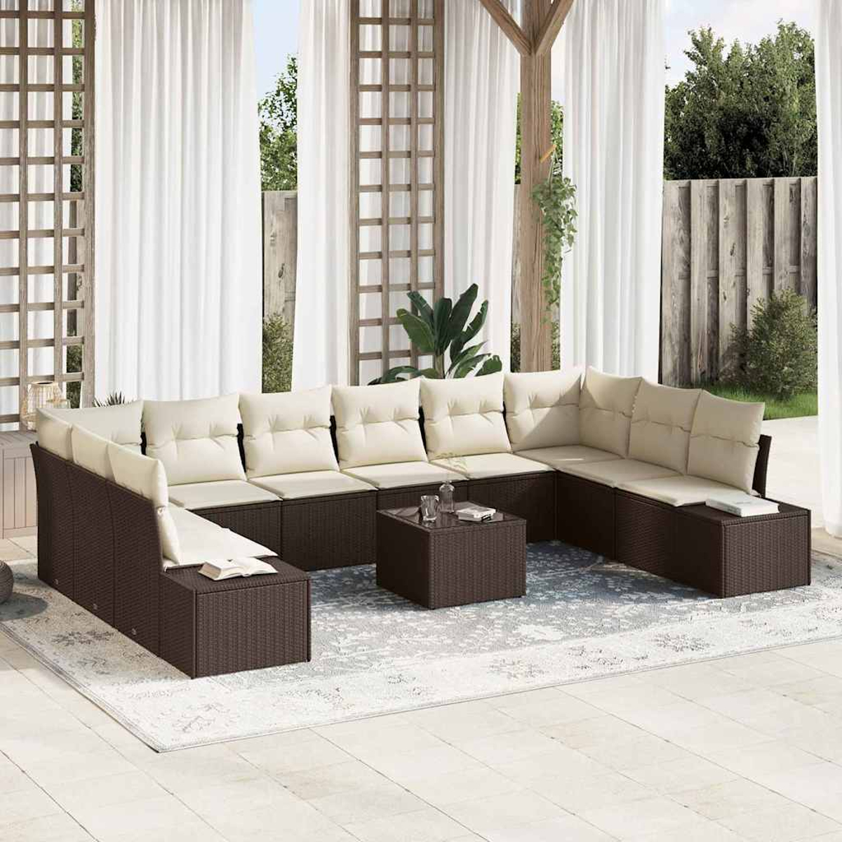 Ein braunes Korbsessel-Ecksofa mit cremefarbenen Kissen auf einem gemusterten Teppich, in einer Terrasse mit weißen Vorhängen und einem kleinen Tisch.