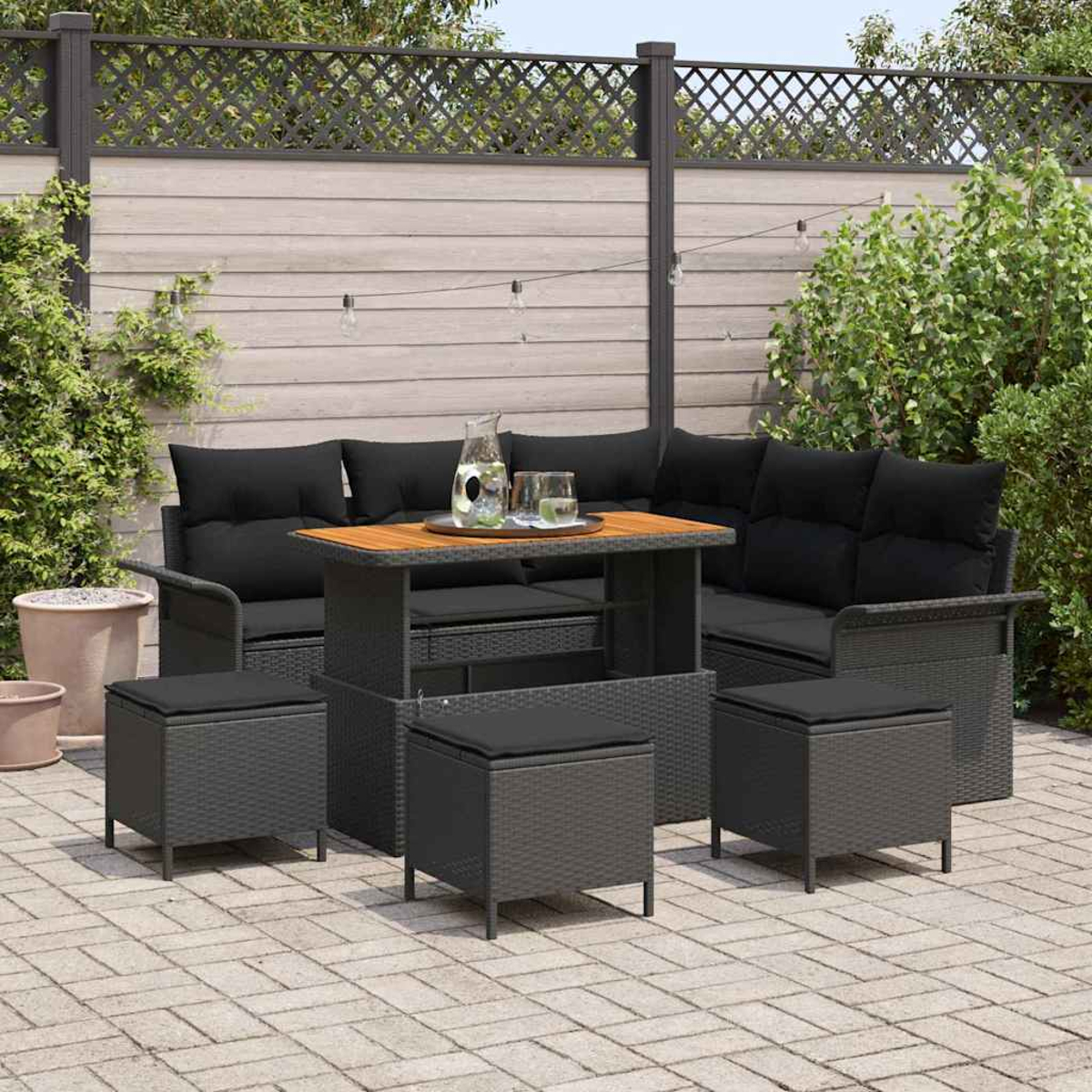 Set di mobili da esterno: divano in vimini grigio con cuscini neri, un tavolo in legno e pouf abbinati su un patio in mattoni.