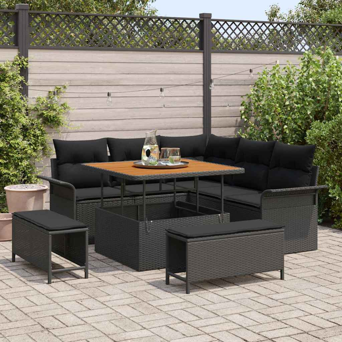 Set da patio con cuscini neri, tavolo in legno e due panche. Su un patio in mattoni. Sfondo: recinzione in legno e vegetazione.