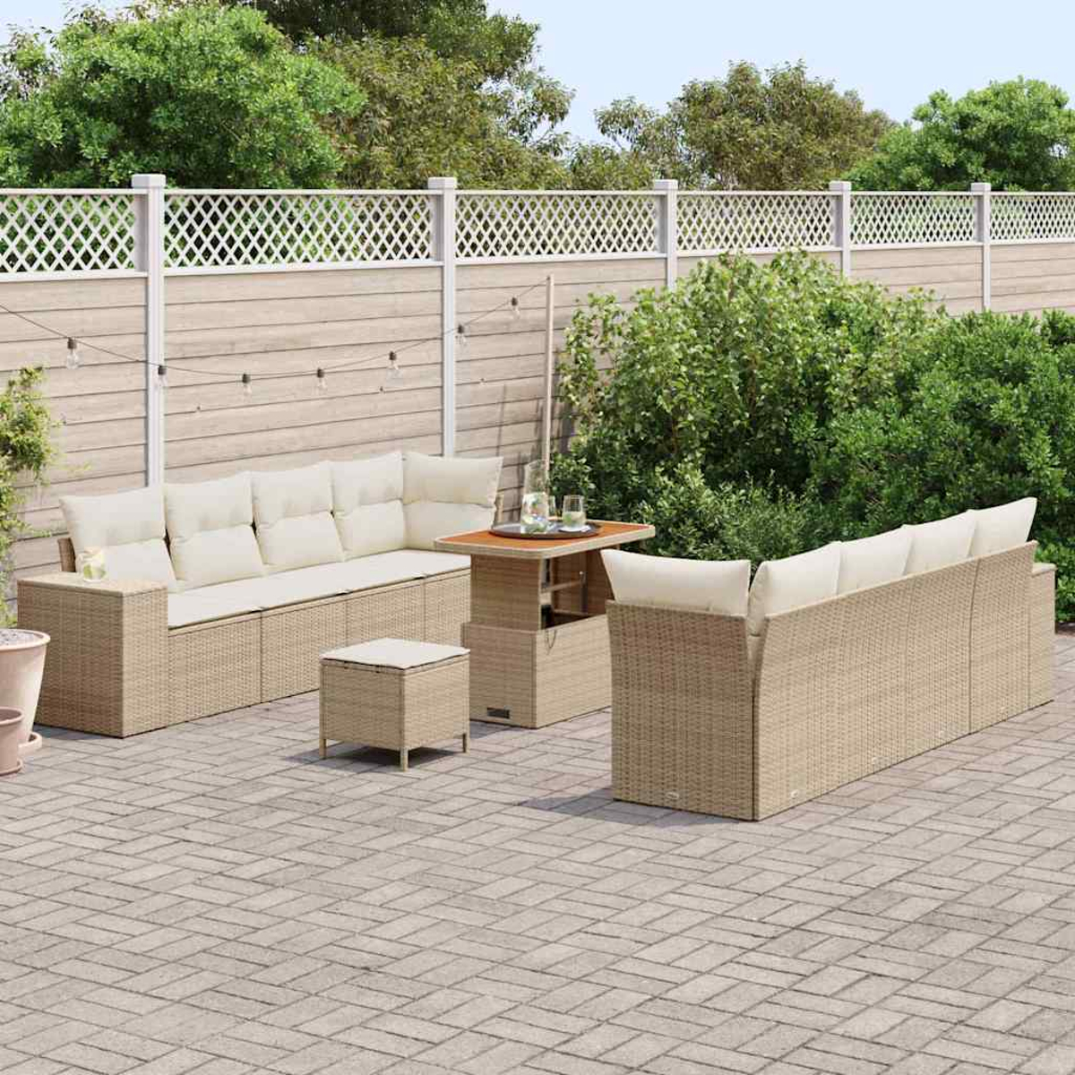Ein beiges Korbsitzgarnitur mit Kissen, ein kleiner Tisch und ein passender Hocker auf einer Backstein-Terrasse, mit Zaun und Grün im Hintergrund.