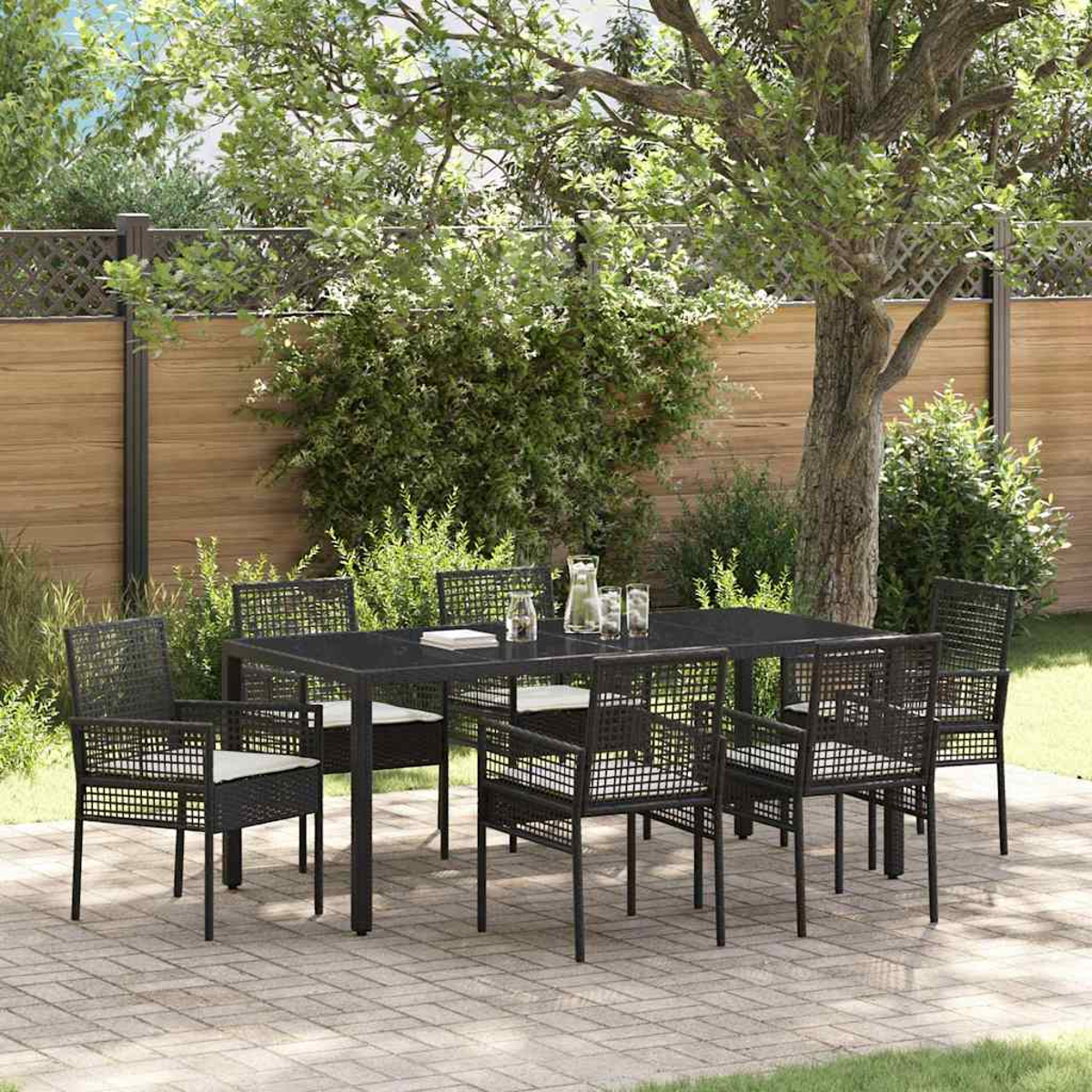 Essgruppe im Freien auf einer Terrasse. Ein dunkler Tisch mit Stühlen und weißen Kissen. Im Hintergrund ein Holzzaun und Bäume.