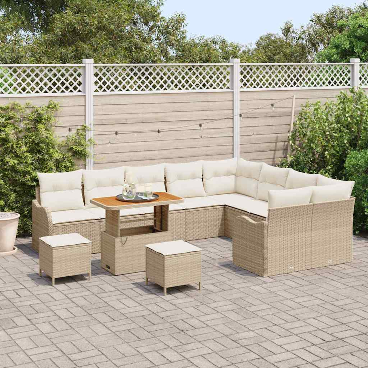 Outdoor-Korbsessel mit cremefarbenen Kissen, Tisch und zwei Hockern. Auf einer Terrasse mit weißem Zaun und Grün im Hintergrund.