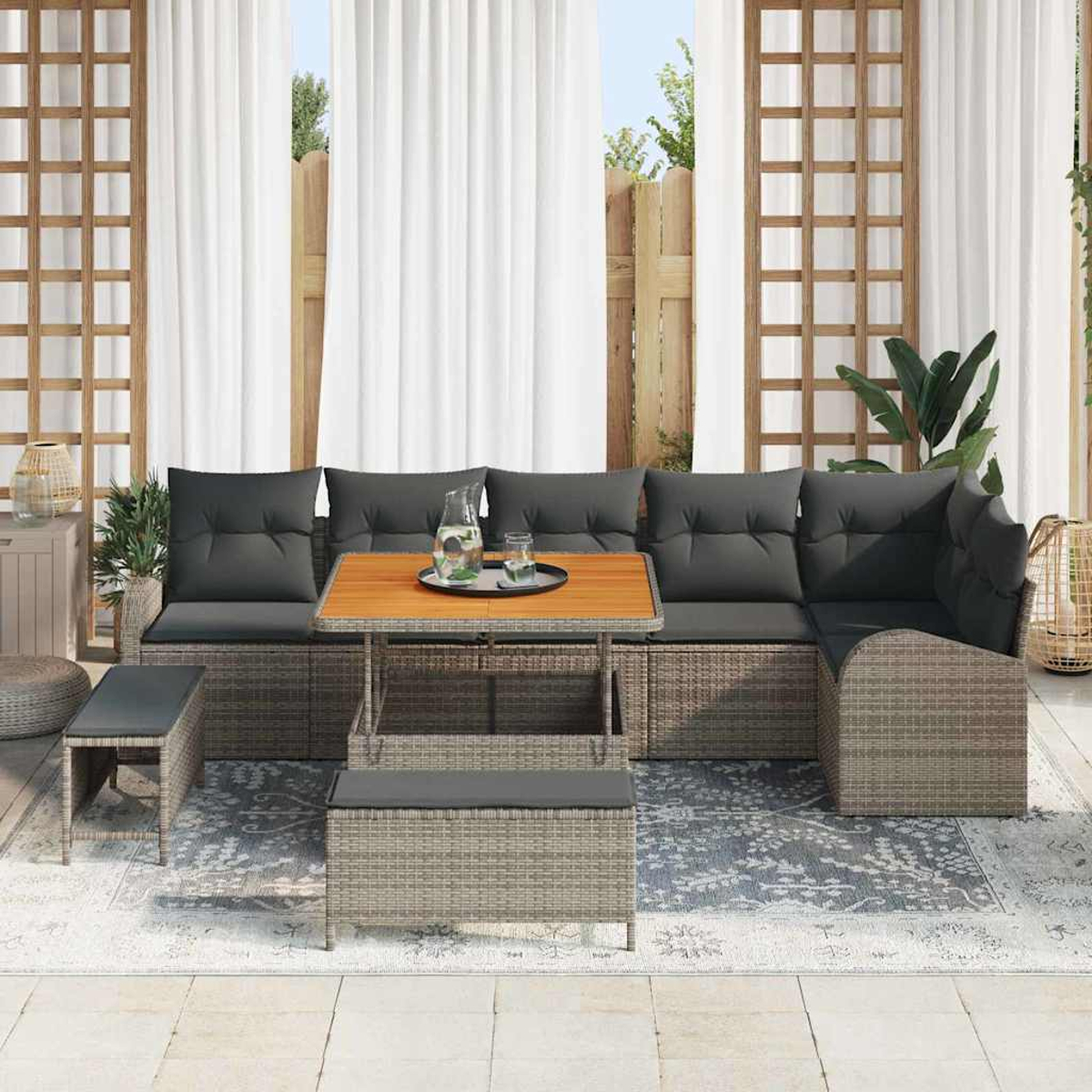 Set di mobili da patio con cuscini grigi e tavolo in legno, su sfondo di tende bianche e recinzione in legno.