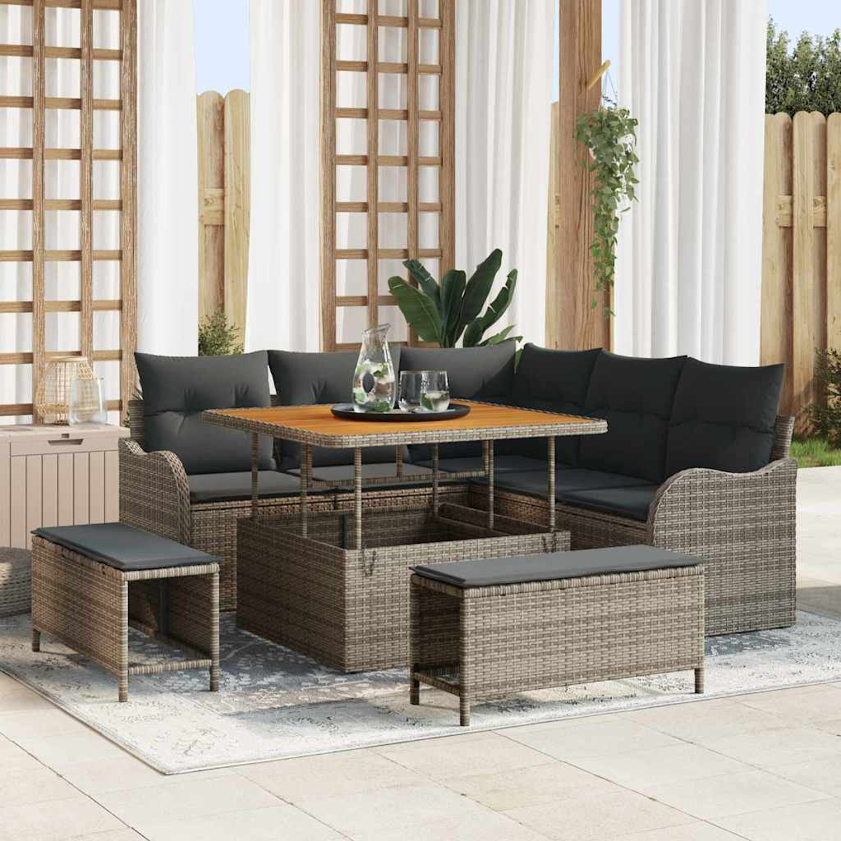 Gartenmöbel-Set mit Tisch, Bänken und Kissen. Weiße Vorhänge und Holzzaun im Hintergrund. Pflanzen sind ebenfalls sichtbar.