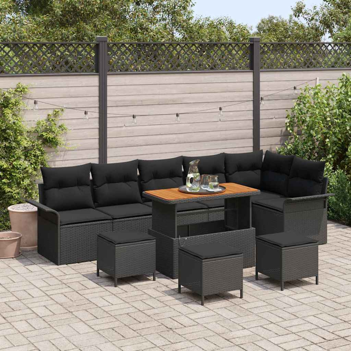Garten-Sitzgruppe mit schwarzen Kissen, einem Tisch und Hockern. Dahinter ein Zaun und Grün.