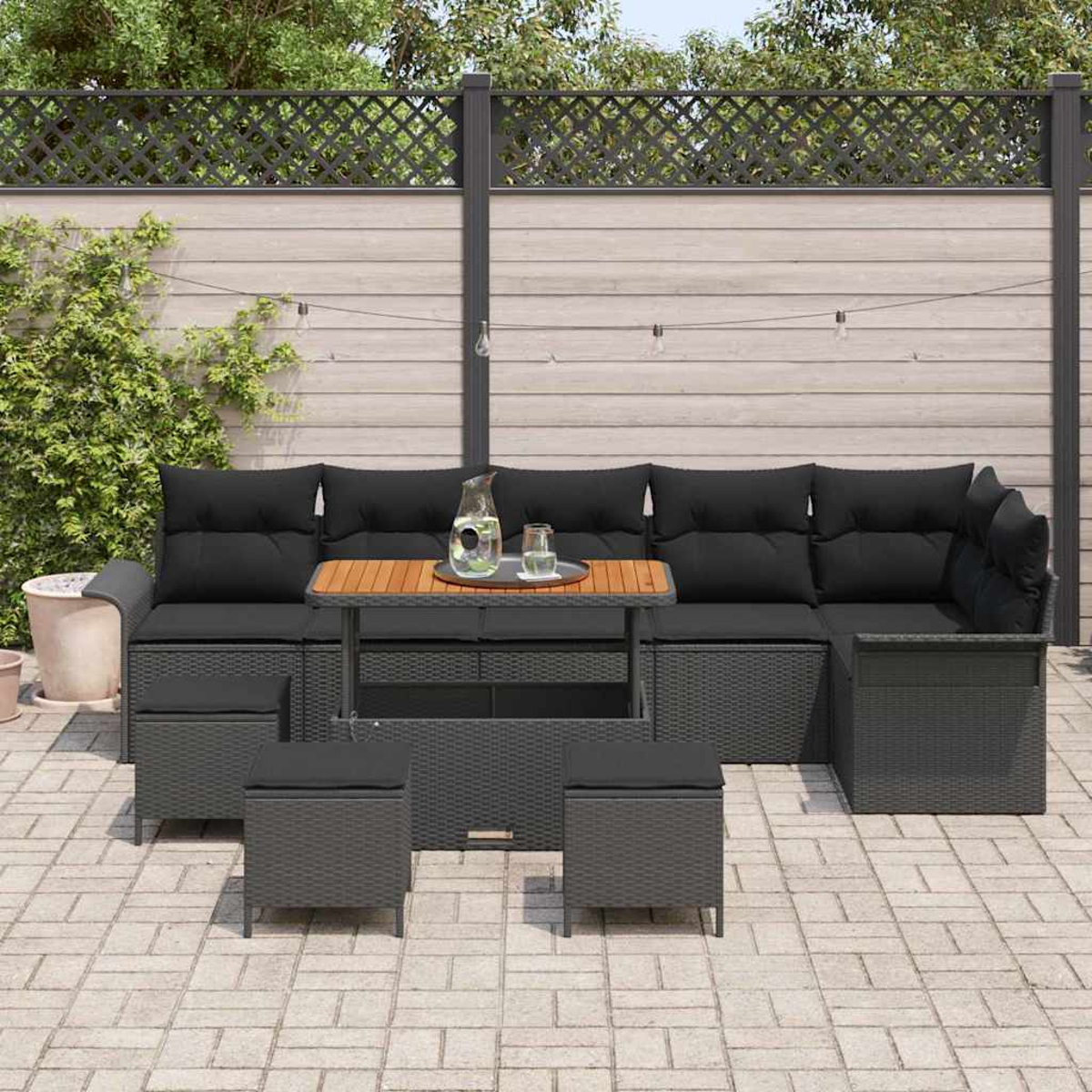 Garten-Terrassenset mit schwarzen Kissen, Tisch und Ottomanen. Ein Holztisch, Getränke und ein Zaun sind sichtbar.