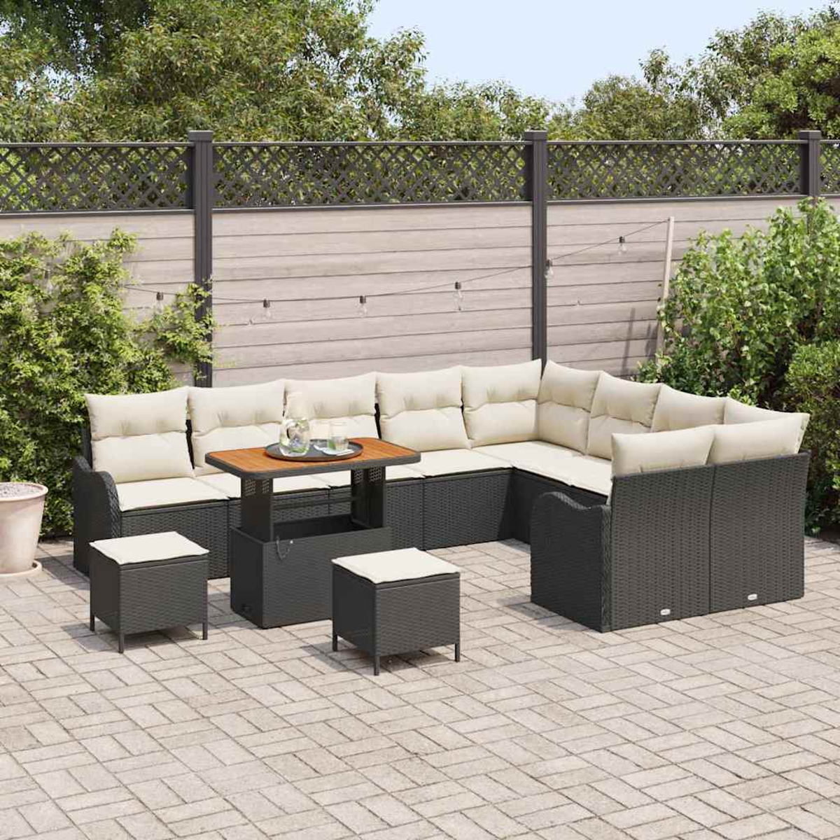 Gartencouch mit beigen Kissen, einem Holztisch und zwei Hockern auf einer gepflasterten Terrasse, mit Zaun und Grün im Hintergrund.