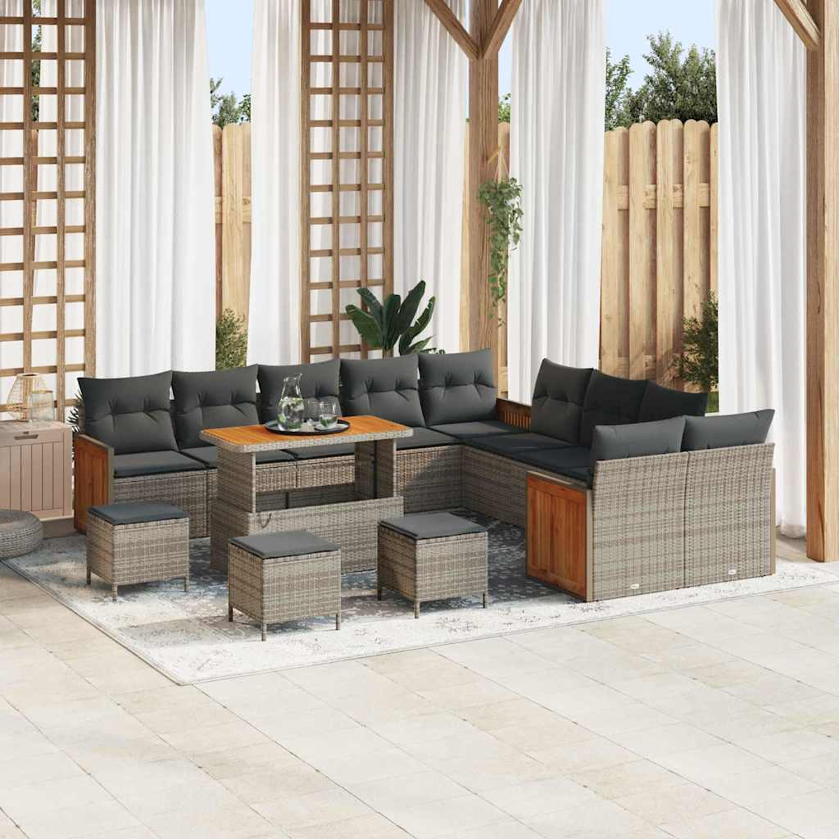 Outdoor-Sitzbereich mit grauen Kissen, einem Holztisch und passenden Hockern auf einem gemusterten Teppich unter einer Holzkonstruktion.