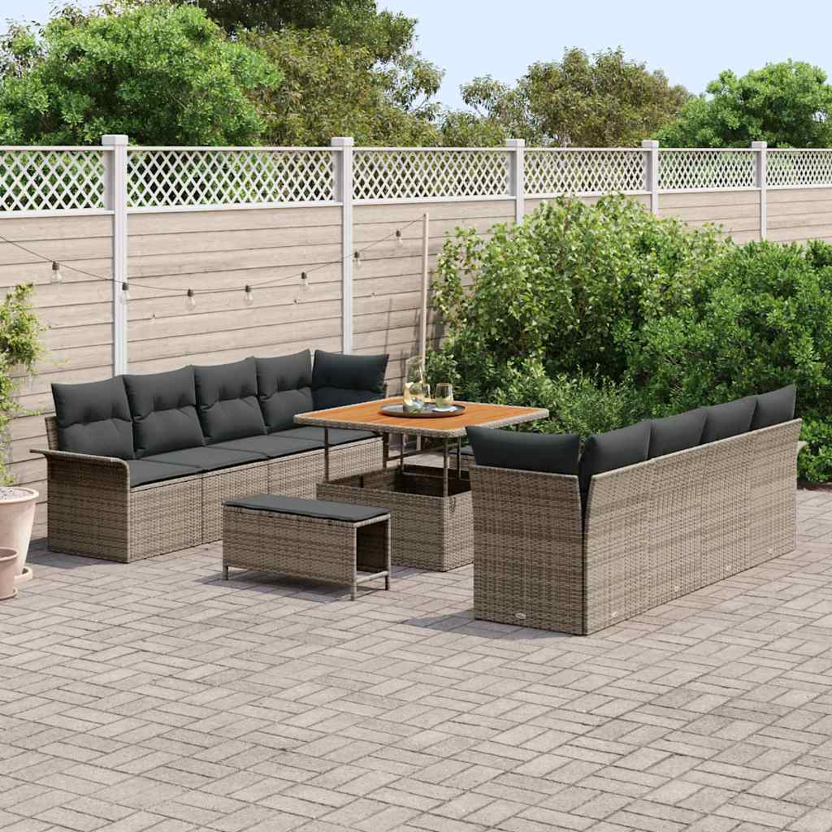 Set patio esterno con divano, tavolo e panca su pavimento pavimentato, circondato da vegetazione e una recinzione bianca.