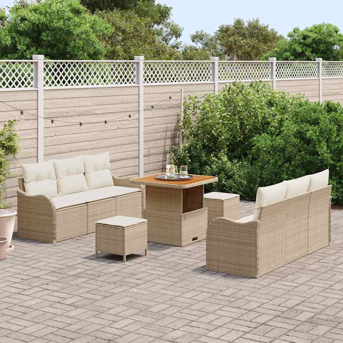 Ein Outdoor-Terrassenset mit gewebten beigen Möbeln, darunter Sofas, ein Tisch und Hocker. Im Hintergrund befinden sich ein Holzzaun und Grün.
