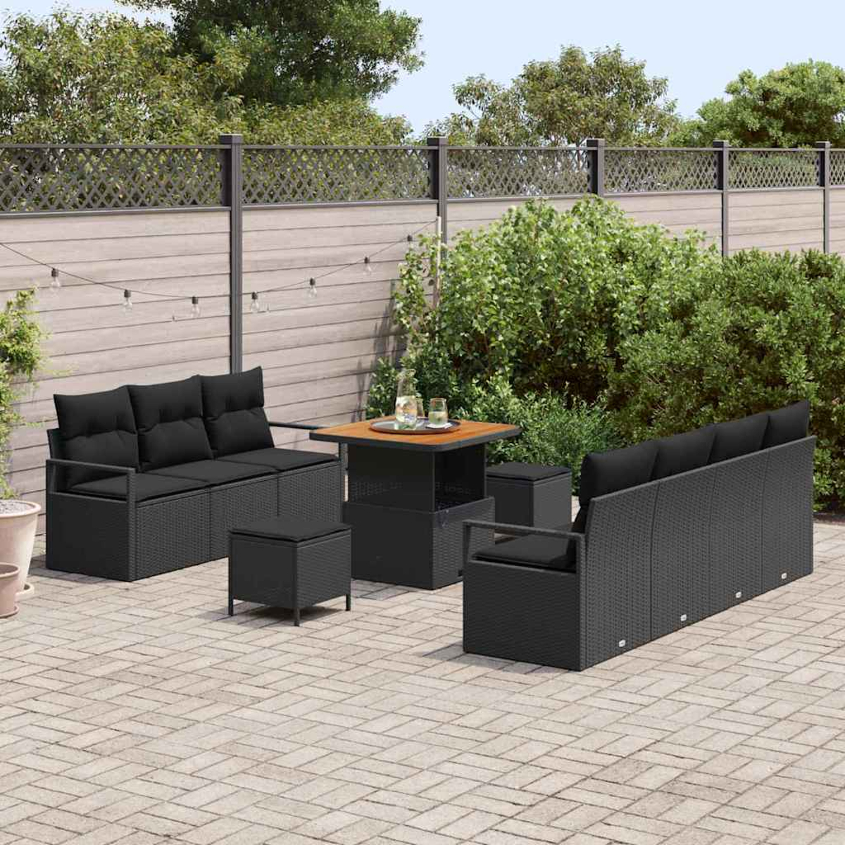Terrassenset mit schwarzen Korbsesseln, Ottomanen und einem Tisch. Die Szene spielt sich auf einer gepflasterten Terrasse mit Zaun und Grün ab.