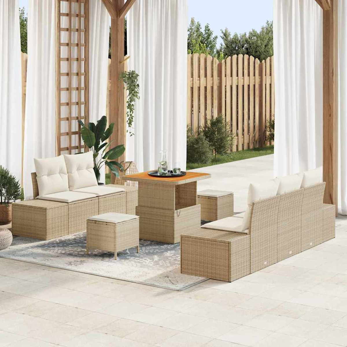 Gartenmöbel-Set mit beigen Korbmöbeln, weißen Kissen und einem Holztisch. Weiße Vorhänge und Holzzaun im Hintergrund.