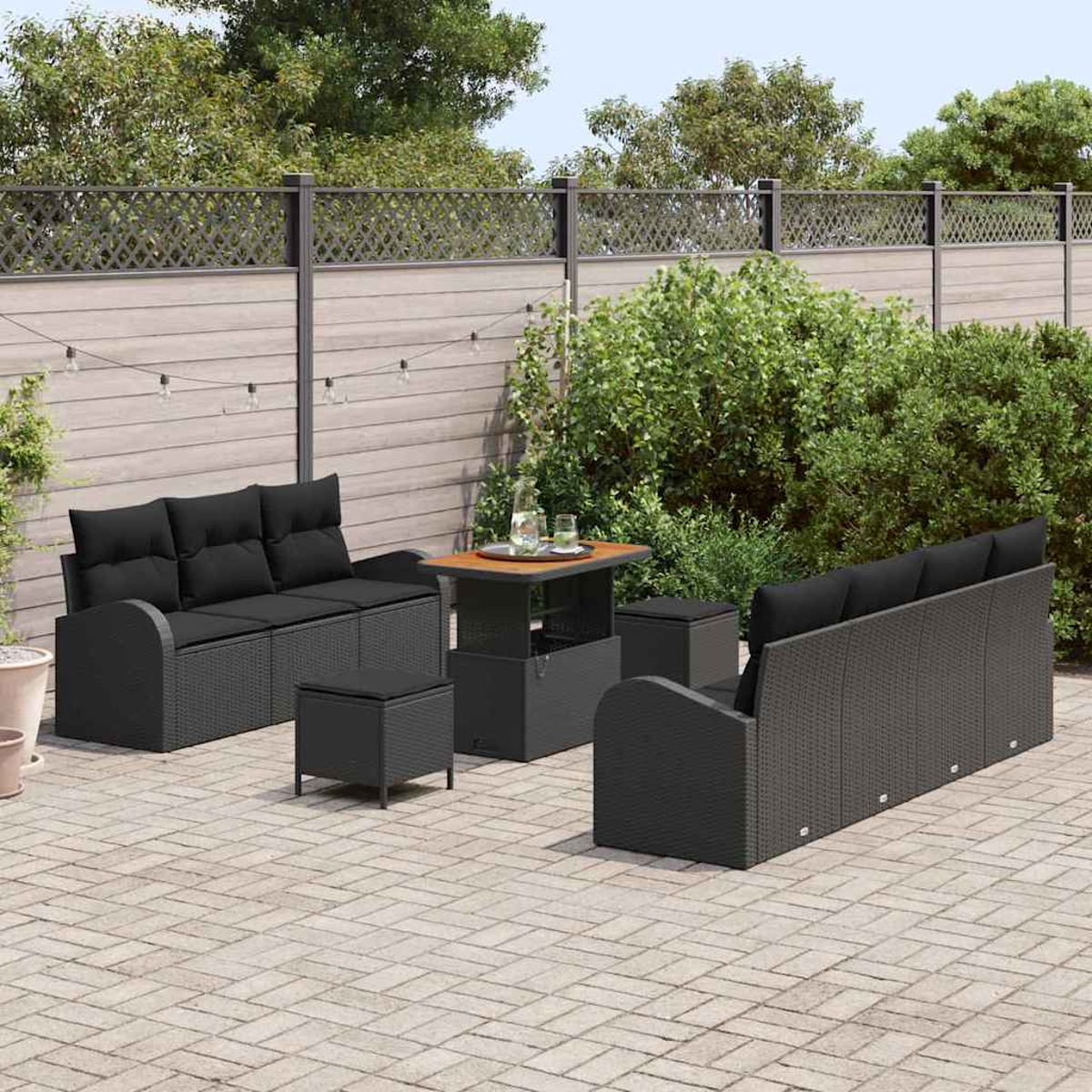 Gartenmöbel-Set auf einer Terrasse. Enthält zwei Sofas, zwei Ottomanen, einen Tisch und einen Beistelltisch. Hintergrund hat einen Zaun und Grün.