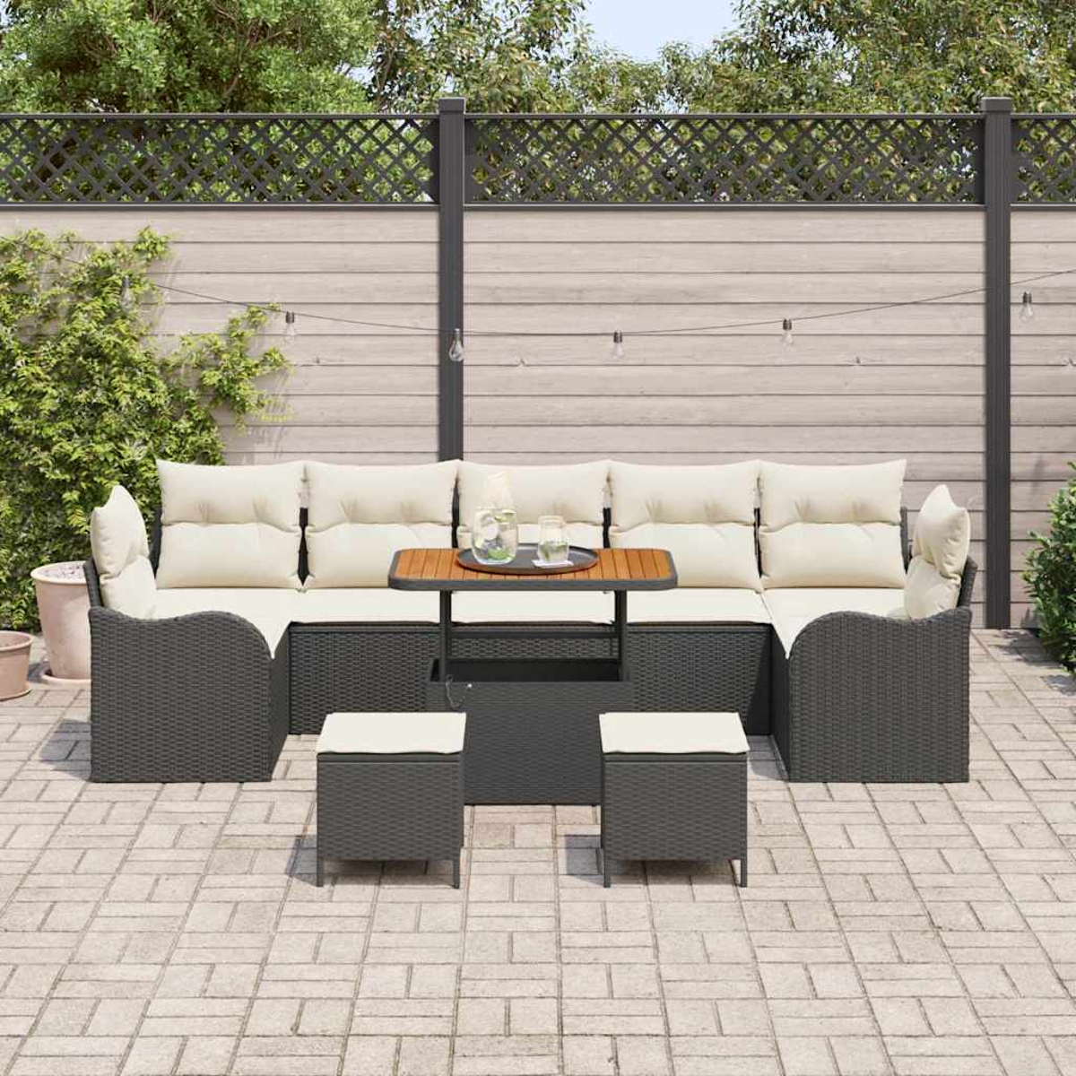 Garten-Sitzgruppe mit Couchtisch und zwei Hockern auf einer Terrasse. Im Hintergrund ein Zaun und Grün.