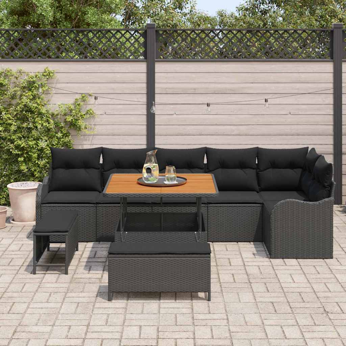 Outdoor-Ecksofa mit schwarzen Kissen und einem Holztisch. Eine Bank und ein kleiner Hocker sind ebenfalls sichtbar. Der Hintergrund hat einen Holzzaun.