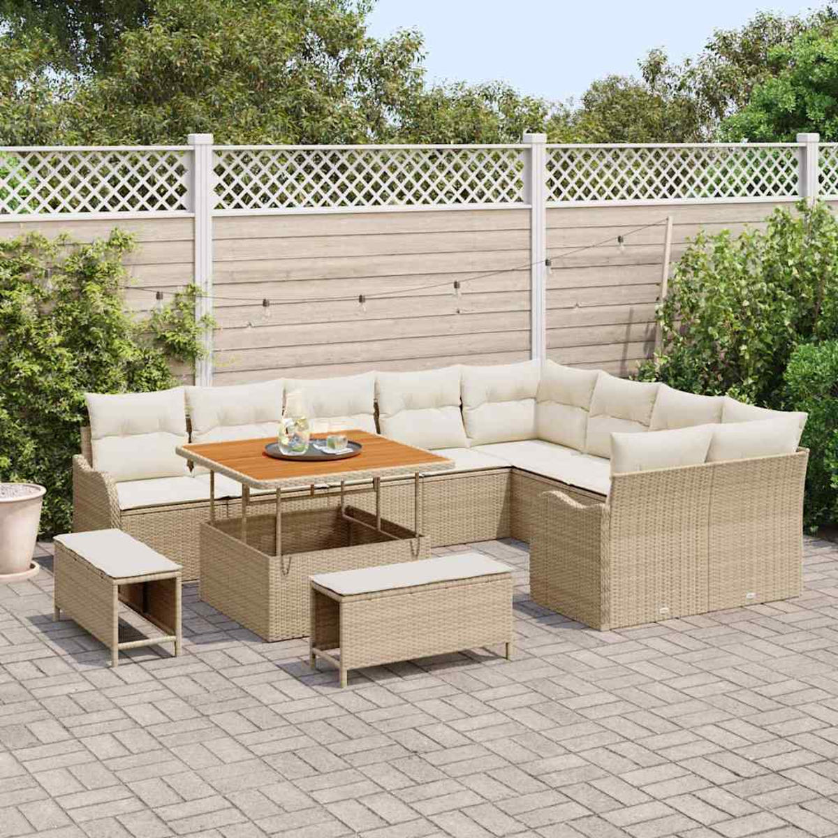 Ein Outdoor-Sofa-Set aus Korbgeflecht mit cremefarbenen Kissen, einem Holztisch und zwei Bänken auf einer Ziegelterrasse vor einem weißen Zaun.