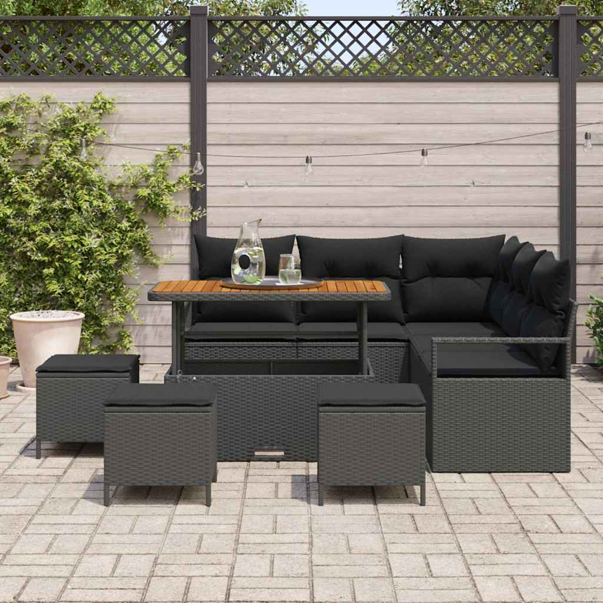 Juego de patio al aire libre con mesa, sofá y taburetes. Cojines negros y mimbre gris. Tapa de mesa de madera. Sobre un patio de piedra, con valla al fondo.