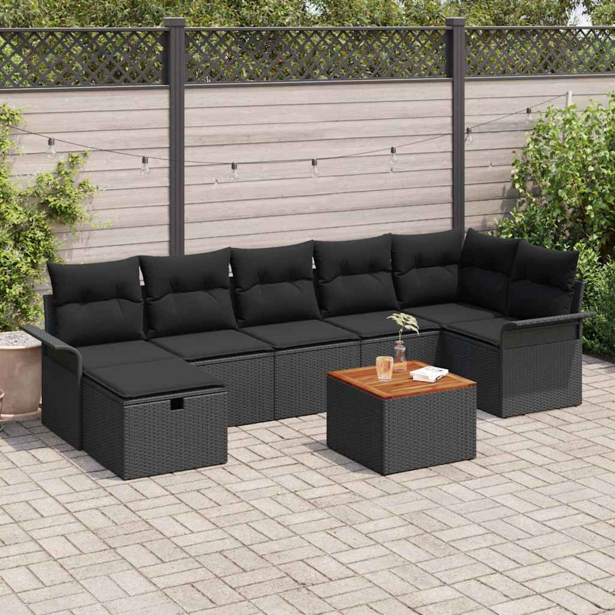 Sofá seccional exterior con cojines negros y una pequeña mesa de madera en un patio de piedra. Valla y plantas al fondo.