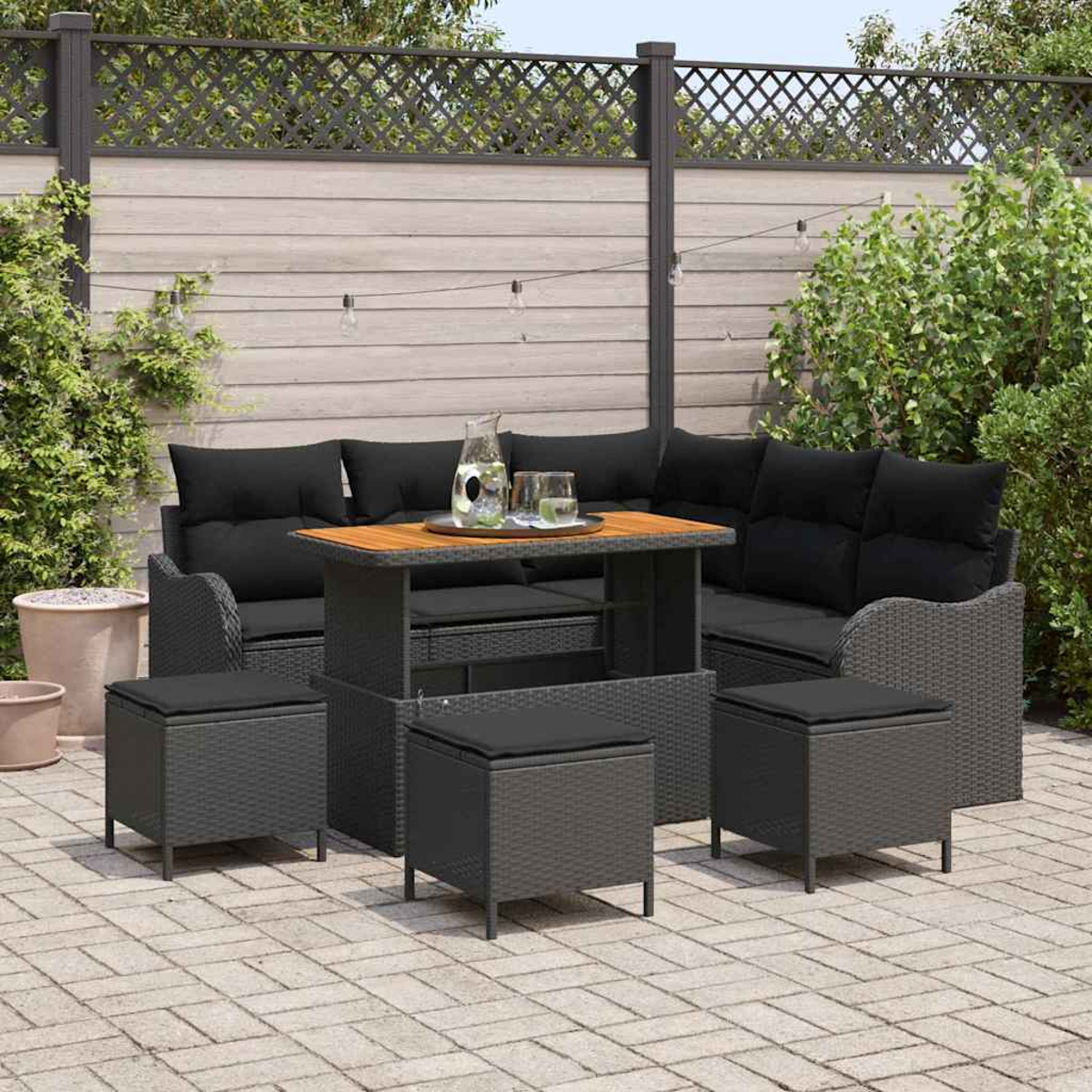 Garten-Terrassenset mit schwarzen Kissen, einem Holztisch und passenden Ottomanen. Vor einem Zaun mit Pflanzen und Lichterketten.