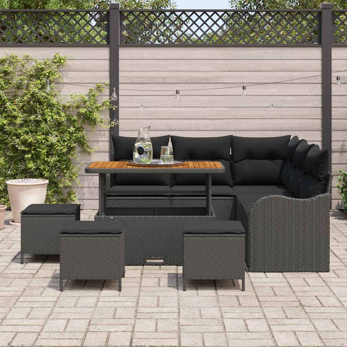 Terrassenset im Freien mit Ecksofa, Tisch und Ottomanen. Schwarze Kissen, graues Weidengeflecht. Holzzaun im Hintergrund.