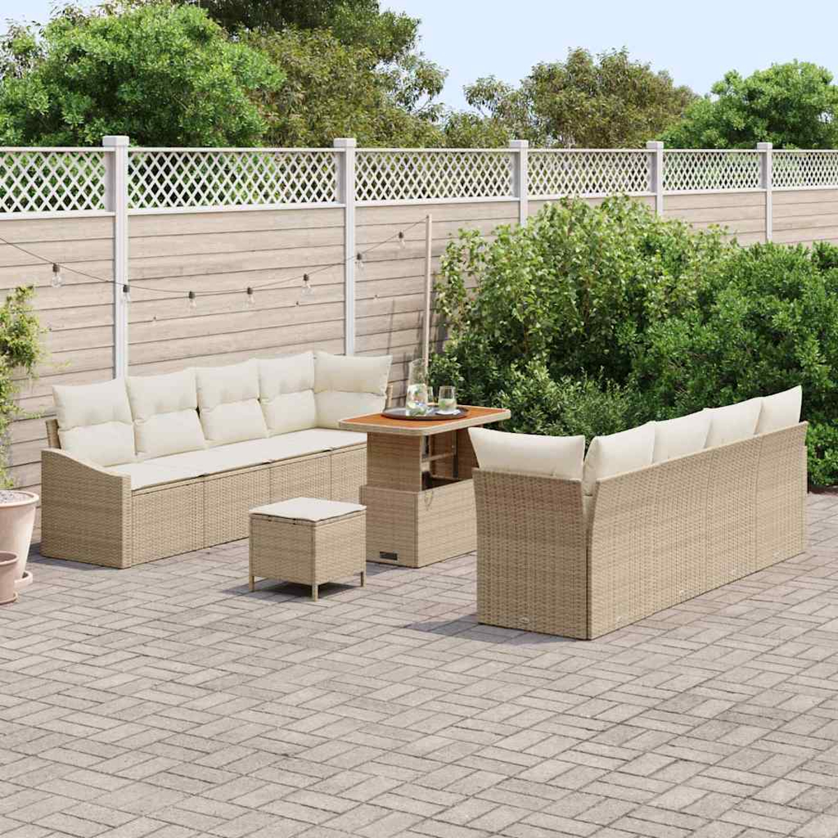 Terrassenset mit beigen Korbsesseln, Tisch und Hocker. Ein weißer Zaun und grüne Pflanzen im Hintergrund.
