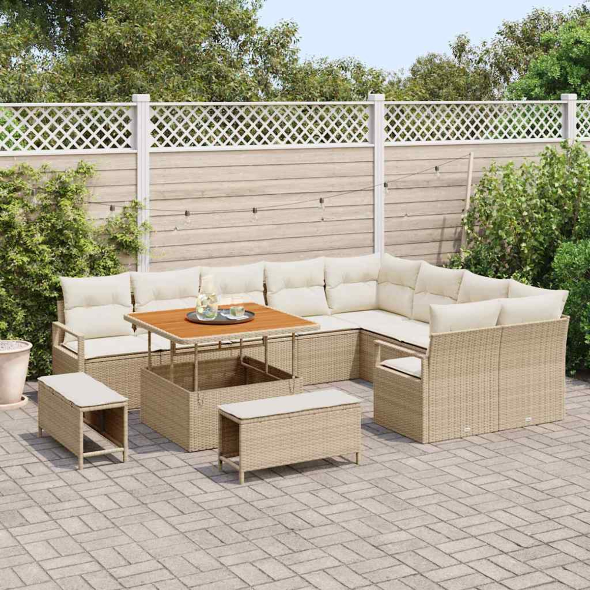 Gartenmöbel-Set. Beigefarbenes Ecksofa aus Korbgeflecht, Couchtisch und Bänke auf einer Stein-Terrasse. Weißer Zaun und Grün im Hintergrund.
