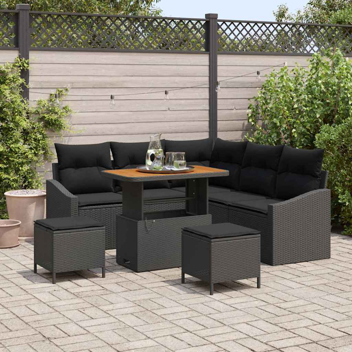 Garten-Sitzgruppe mit schwarzen Kissen, Tisch und Hockern auf einer Terrasse. Im Hintergrund sind ein Zaun und Pflanzen zu sehen.